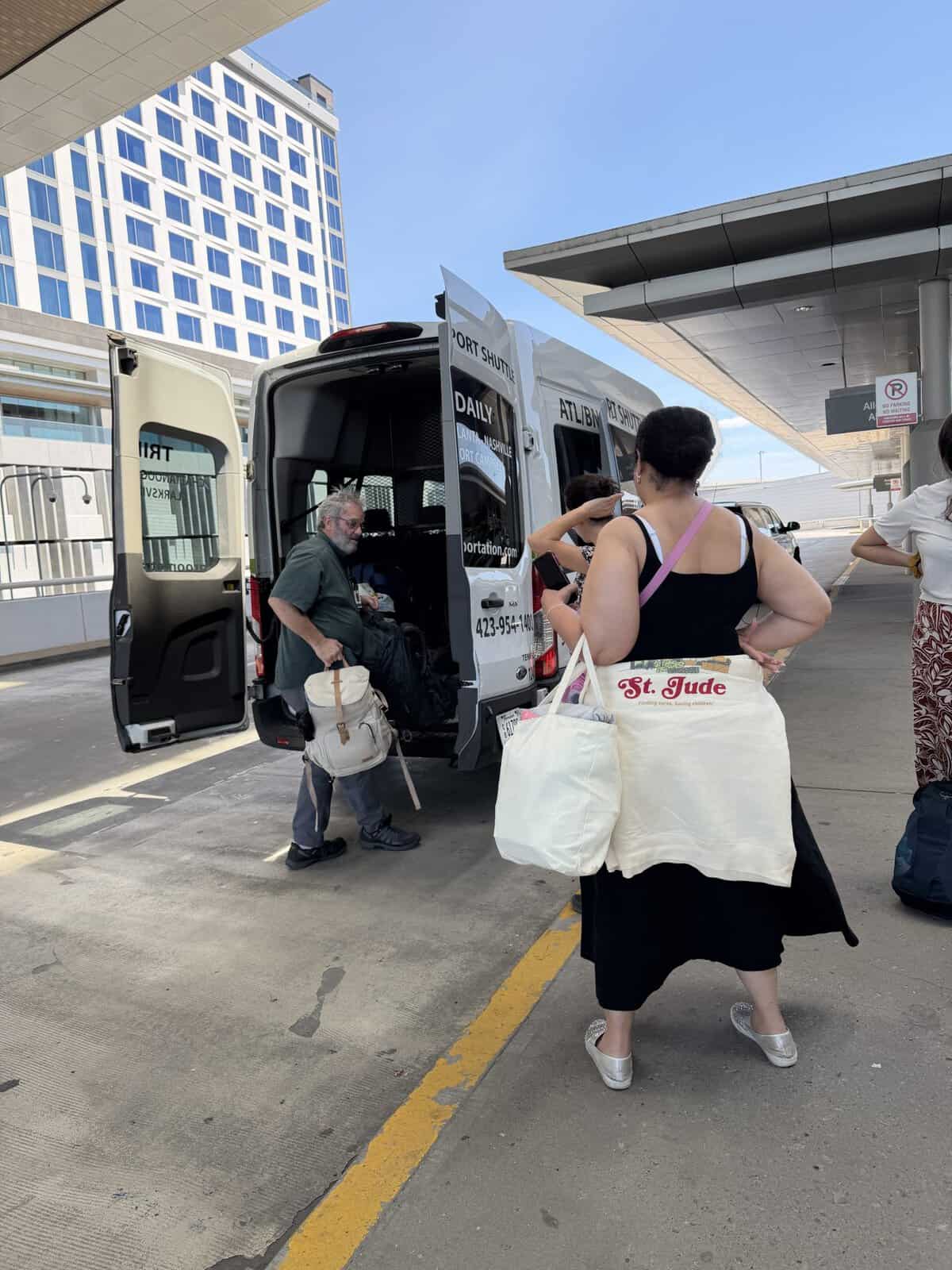 Passengers load luggage into a white airport shuttle van with open doors at a curbside drop-off under a modern overhang, with a tall building in the background.