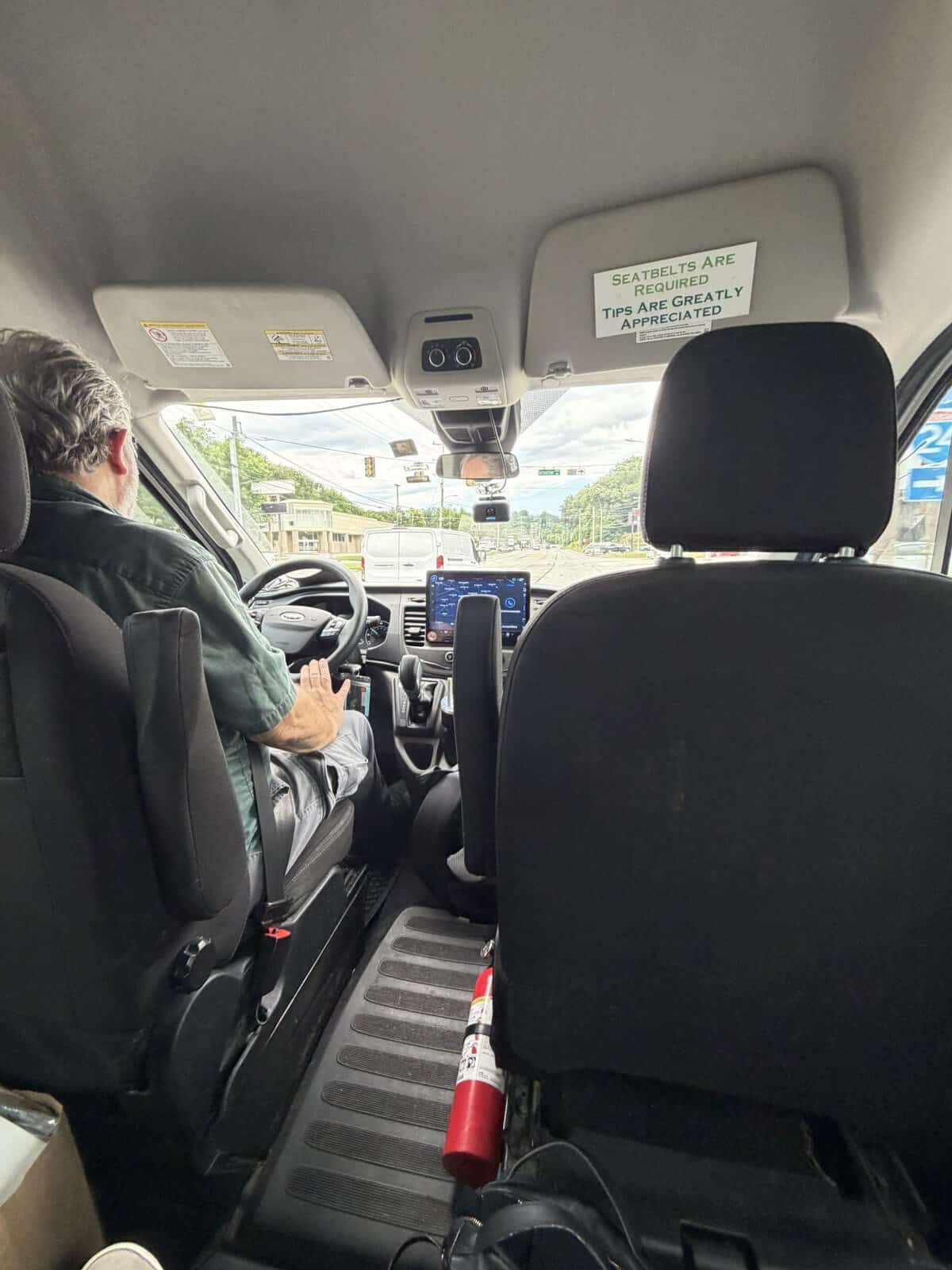 Driver in a green shirt sits at the wheel of a moving van, with a touchscreen dashboard visible and a red fire extinguisher on the floor near the front seat.