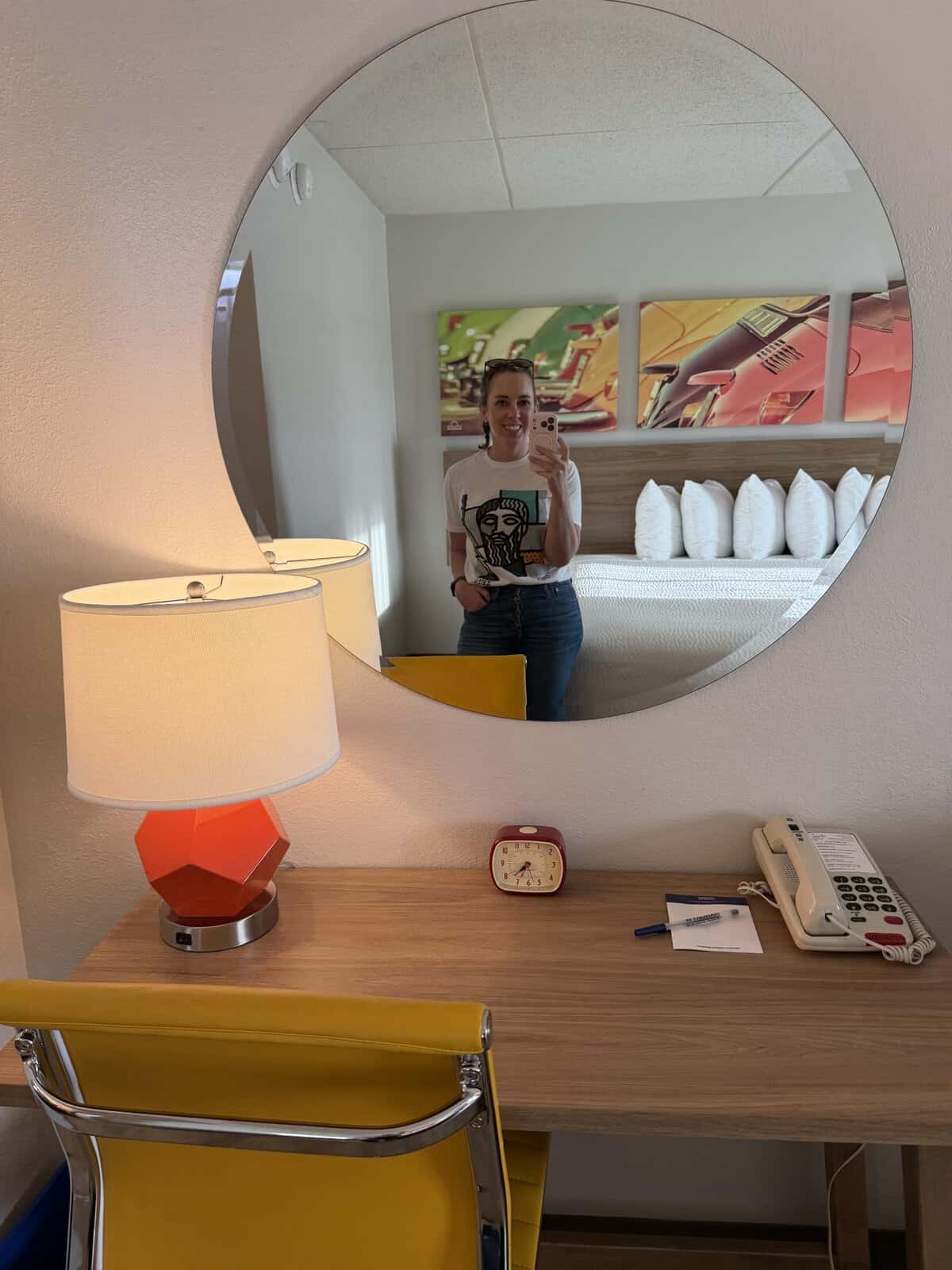 Carli standing in front of the mirror selfie in a hotel room workspace; round wall mirror over a desk with a yellow chair, orange geometric lamp, clock, and phone nearby.