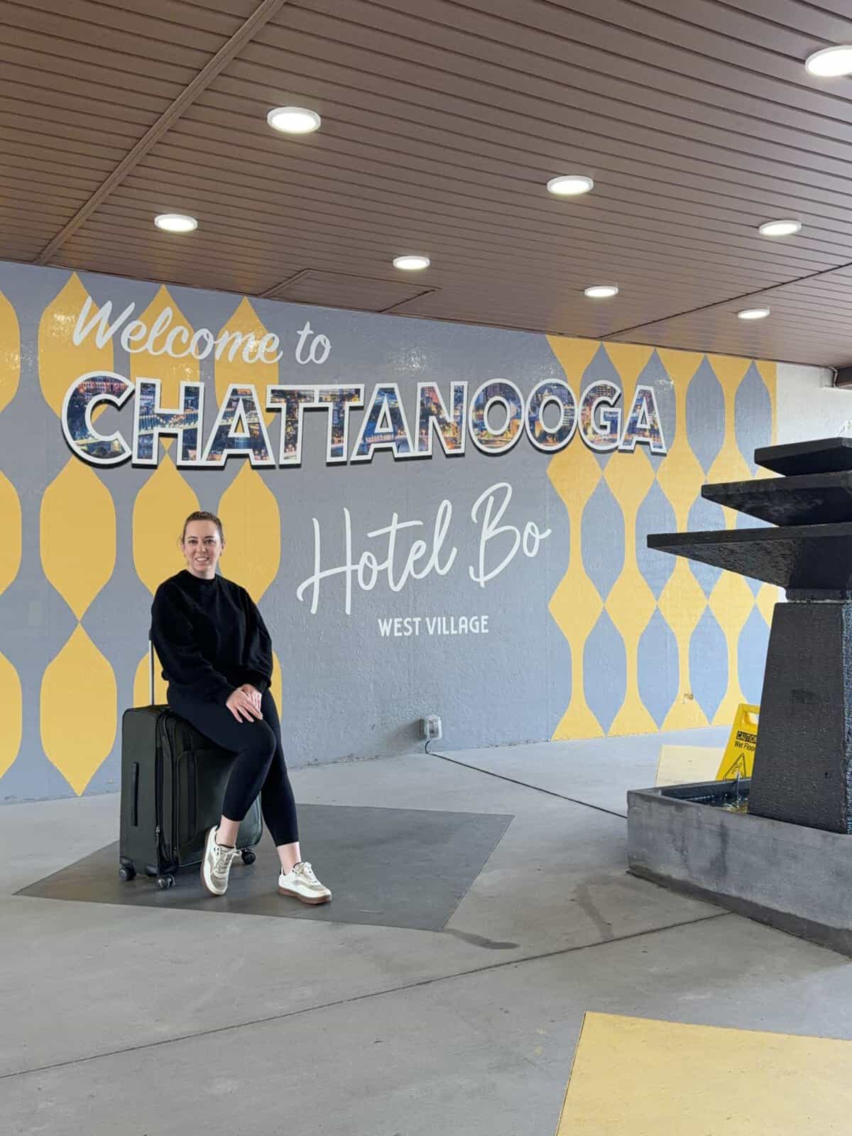 Carli sitting on a rolling suitcase in front of a Chattanooga hotel mural reading'Welcome to Chattanooga' and 'Hotel Bo West Village.'