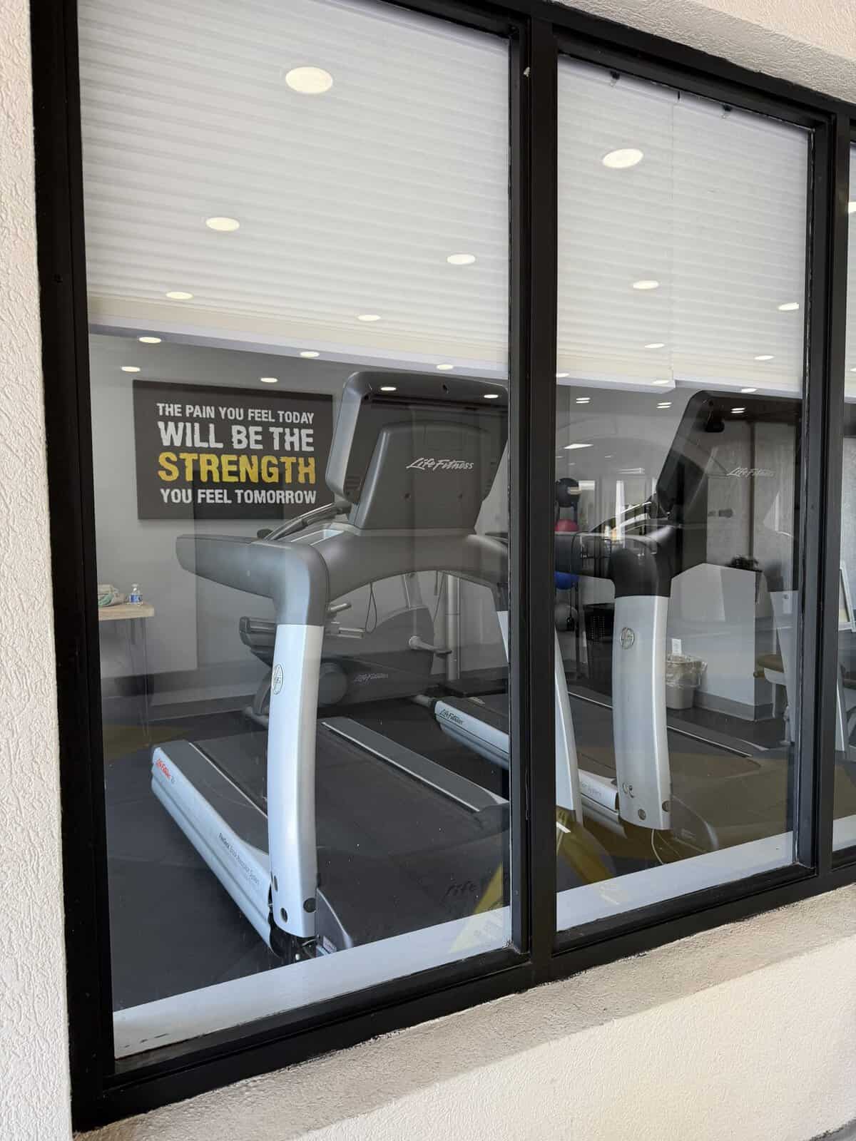 Gym interior seen through a window with treadmill machines and a motivational poster reading,'The pain you feel today will be the strength you feel tomorrow.'