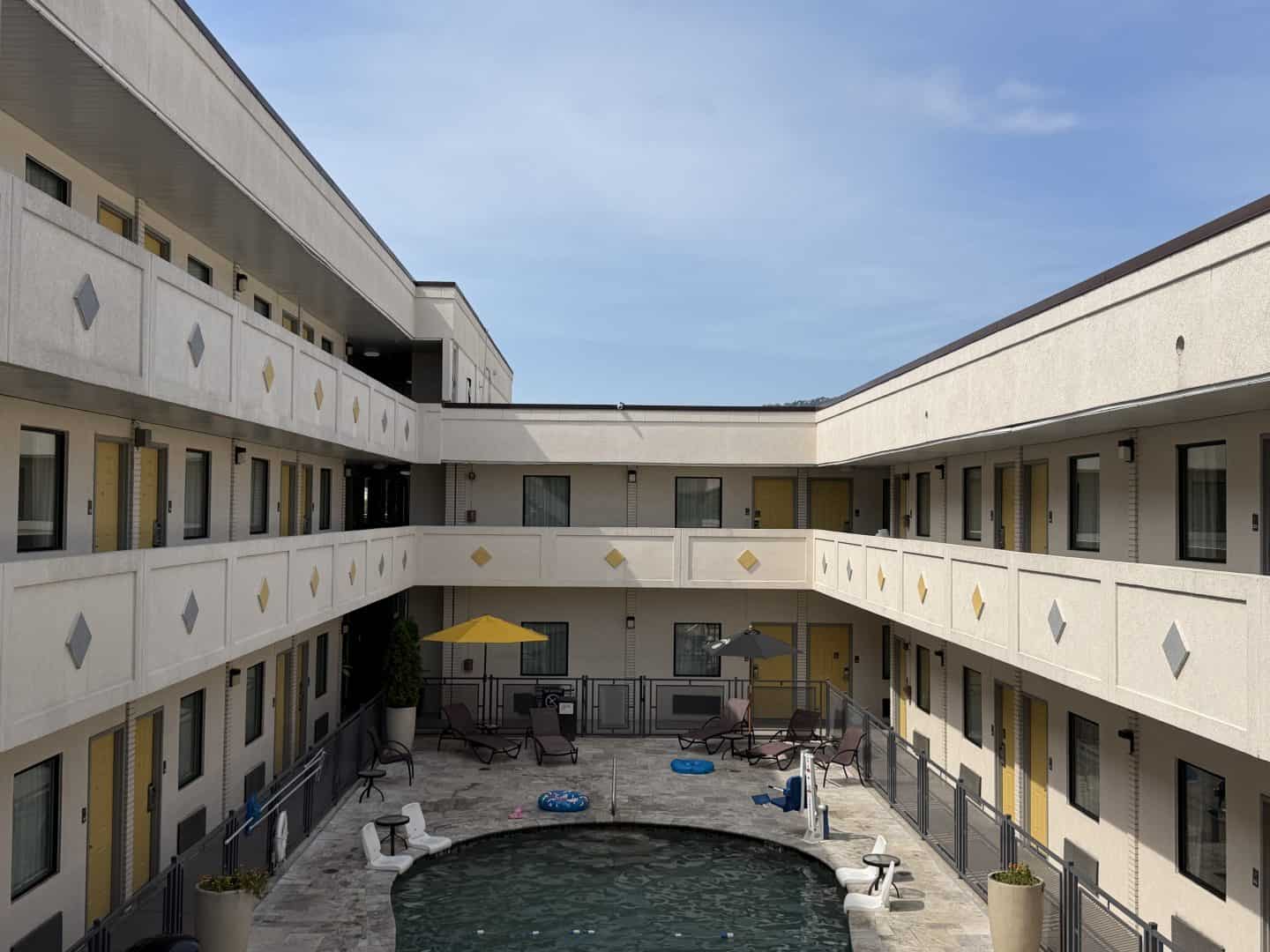 Courtyard scene between beige apartment buildings with a central pool, lounge chairs, and yellow doors under a blue sky.