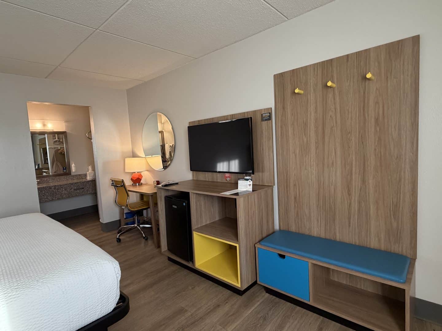 Hotel Bo chattanooga room interior with a bed on the left, a wood desk, wall-mounted TV, oval mirror, and colorful storage cubbies in yellow and blue beneath a wood-paneled wardrobe with three yellow hooks.