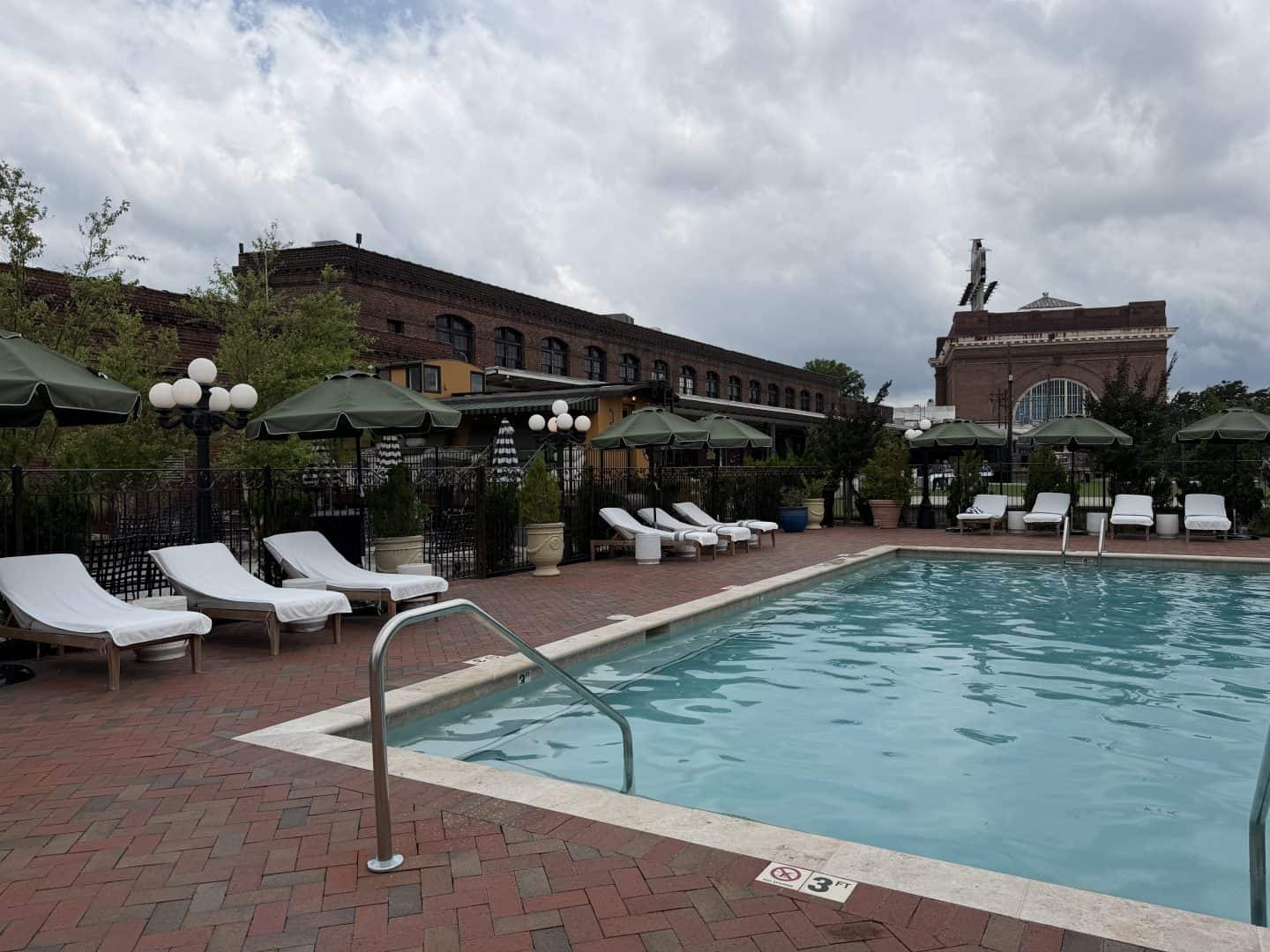 Hotel Chalet Poolside area with turquoise water, white lounge chairs, and green umbrellas against brick buildings under a cloudy sky (outdoor leisure space).