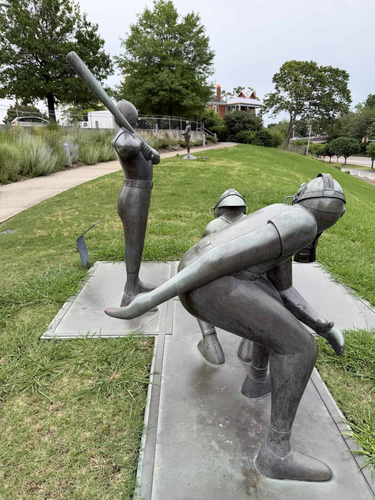 Metal sculpture of three baseball players in action on a grassy park, with one batter swinging and another sliding, mounted on metal bases on a lawn.