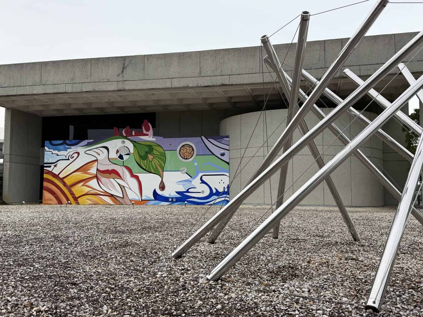Outdoor scene with a colorful graffiti mural on a gray concrete wall under a building, and a cluster of shiny stainless steel poles arranged diagonally in the foreground.