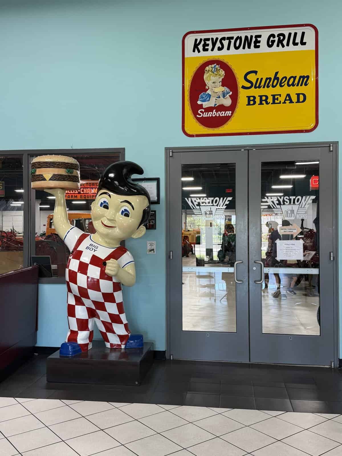 keystone_grill_bobs_big_boy_statue - Adventures with Carli Inside a retro-looking space with a large Keystone Grill Sunbeam Bread sign on a turquoise wall and a Big Boy statue holding a hamburger near glass doors.