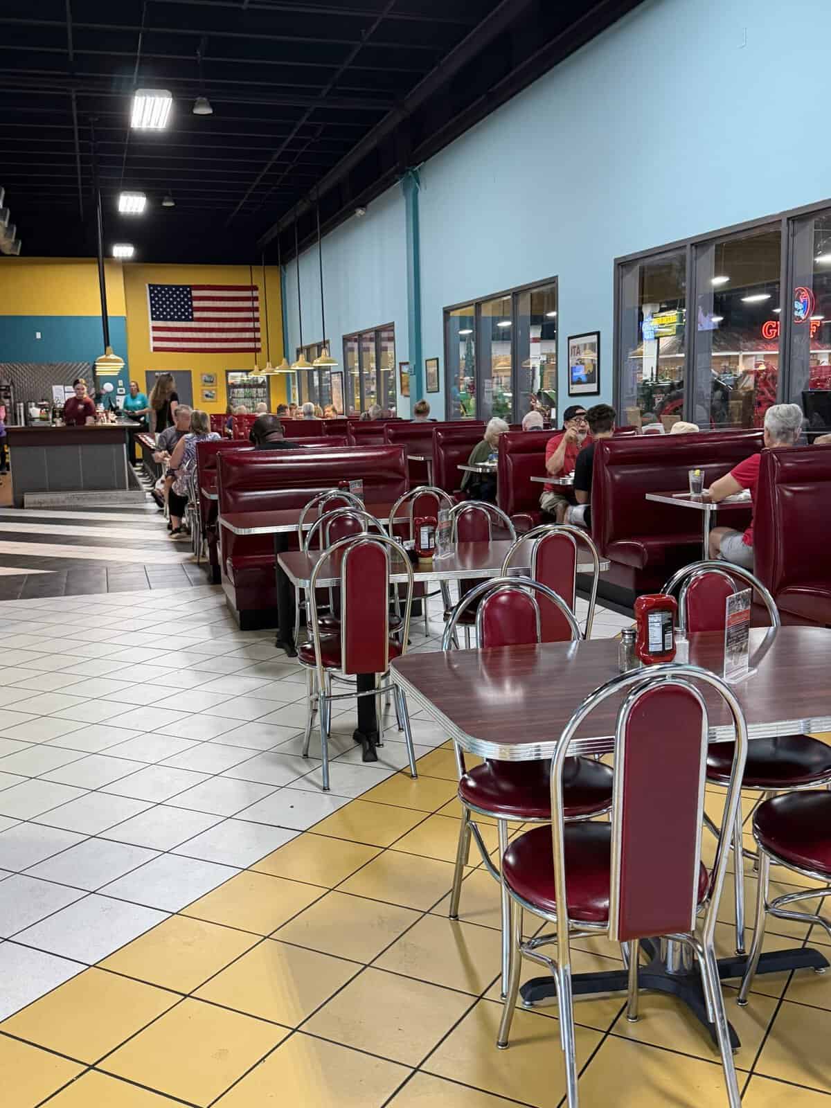keystone_grill_interior - Adventures with Carli Retro diner interior with red vinyl booths, chrome tables, checkered tile floor, and an American flag on a yellow wall; customers sit and eat.