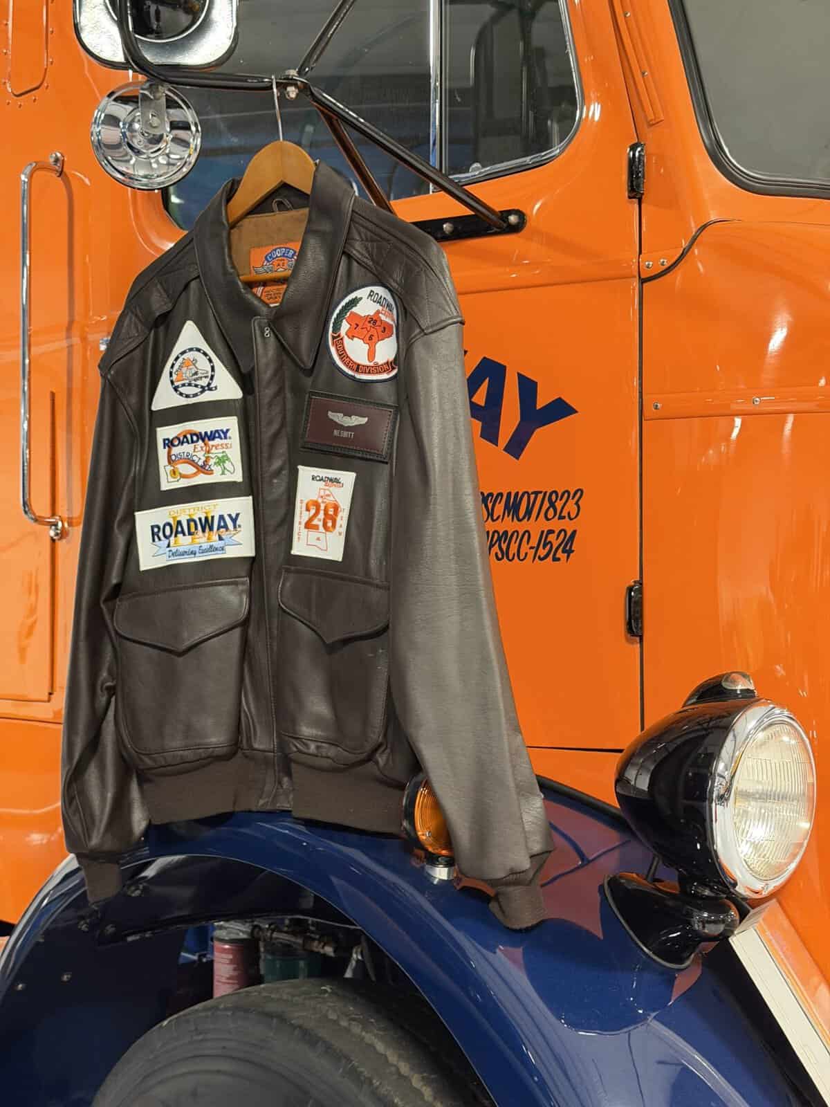 keystone_museum_roadway_jacket - Adventures with Carli brown leather bomber jacket with patches displayed in front of blue and orange truck