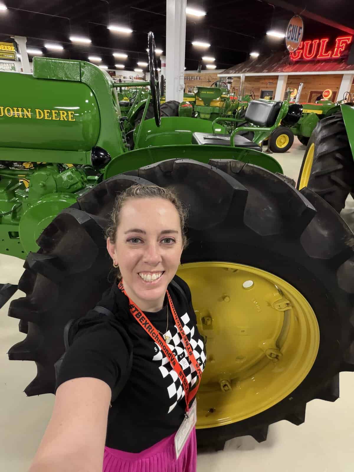 keystone_tractor_museum_carli - Adventures with Carli Selfie of Carli in a black shirt with a red lanyard, standing in front of a large green John Deere tractor with a yellow wheel behind her inside a showroom.