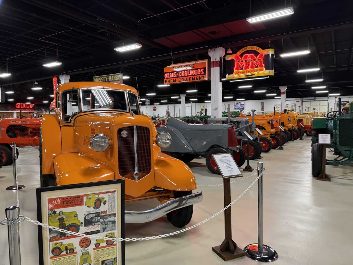 keystone_tractor_museum_minneapolis_moline - Adventures with Carli Front-facing orange vintage tractor in a museum display with retro farm equipment signs and a row of other tractors in the background, under bright ceiling lights.