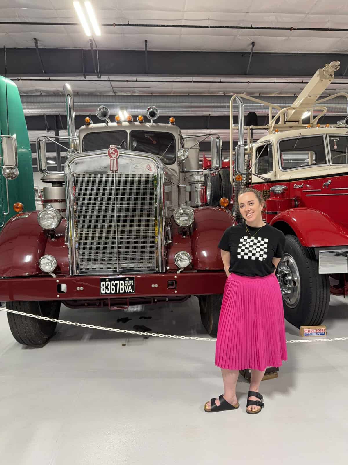 keystone_truck_museum_carli - Adventures with Carli Carli dressed in a black shirt with a white checkerboard design and a bright pink pleated skirt posing in front of a vintage red truck inside a museum-like showroom.