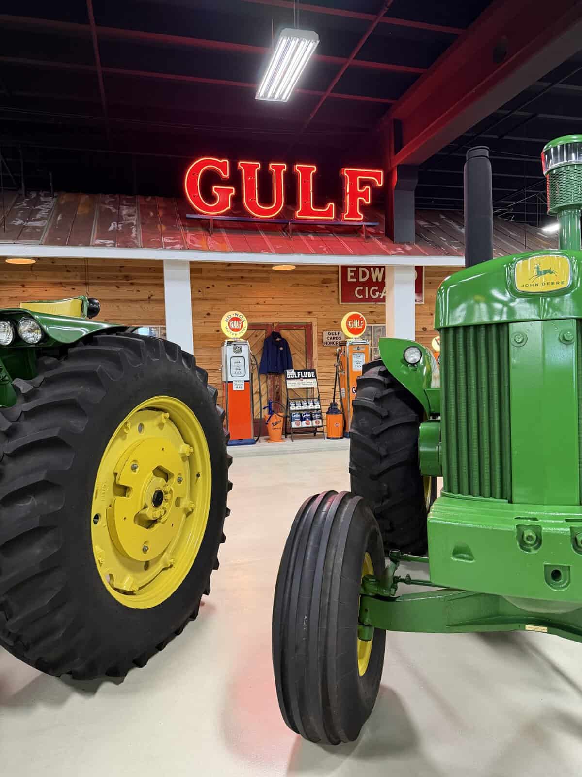 keystone_truck_museum_golf_gas_station - Adventures with Carli Indoor museum or showroom featuring vintage tractors; prominent green John Deere tractor with yellow wheels in the foreground and a large red Gulf neon sign in the background.