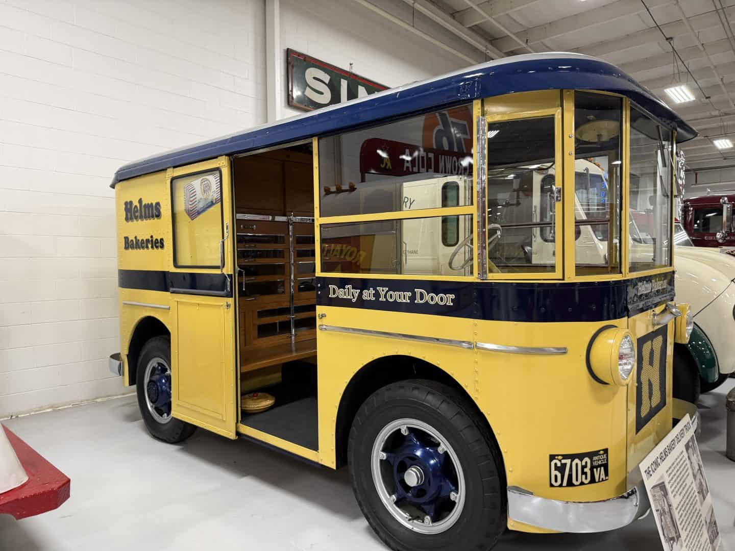 keystone_truck_museum_helms_Bakery - Adventures with Carli Front and side view of a yellow and blue vintage Helms Bakeries delivery van with its passenger doorway open inside a museum showroom.