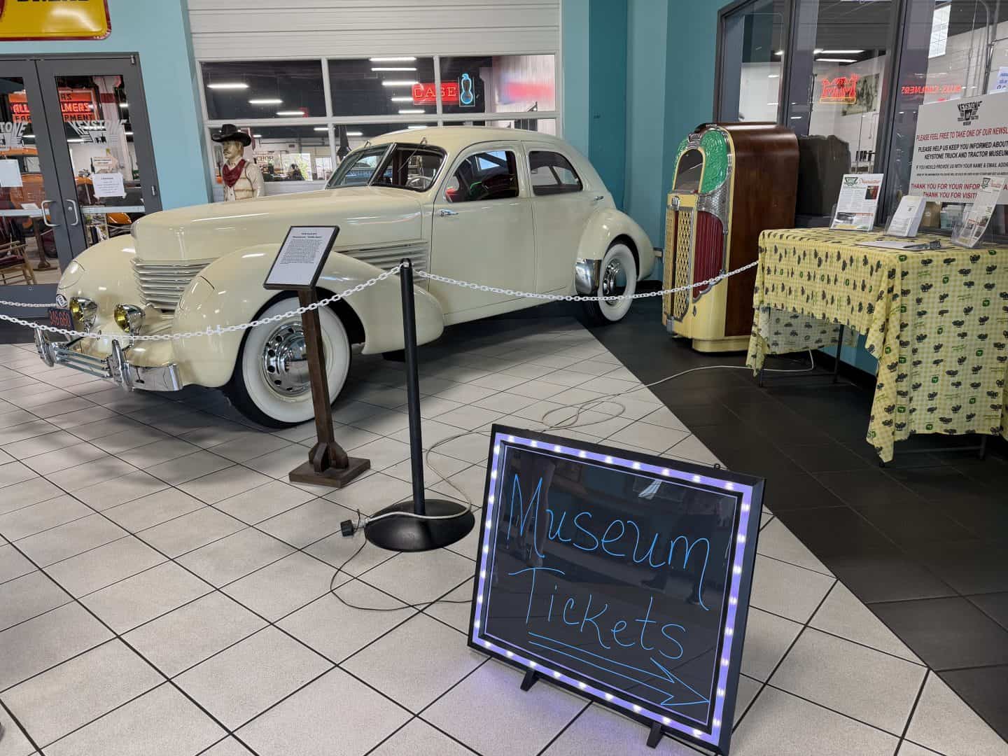 keystone_truck_museum_tickets - Adventures with Carli Cream-colored vintage car on display behind a rope barrier in a showroom; foreground shows a chalkboard sign reading'Museum Tickets'.