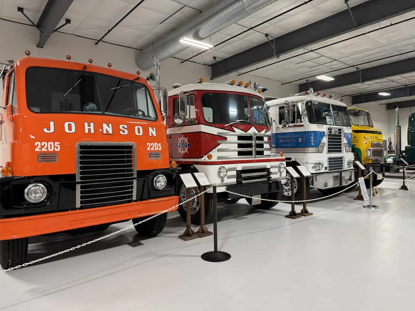 keystone_truck_museum_transformer_trucks - Adventures with Carli Row of four vintage semi trucks in a museum showroom, starting with an orange Johnson truck, black and white truck, then white with blue, ending in a yellow truck.
