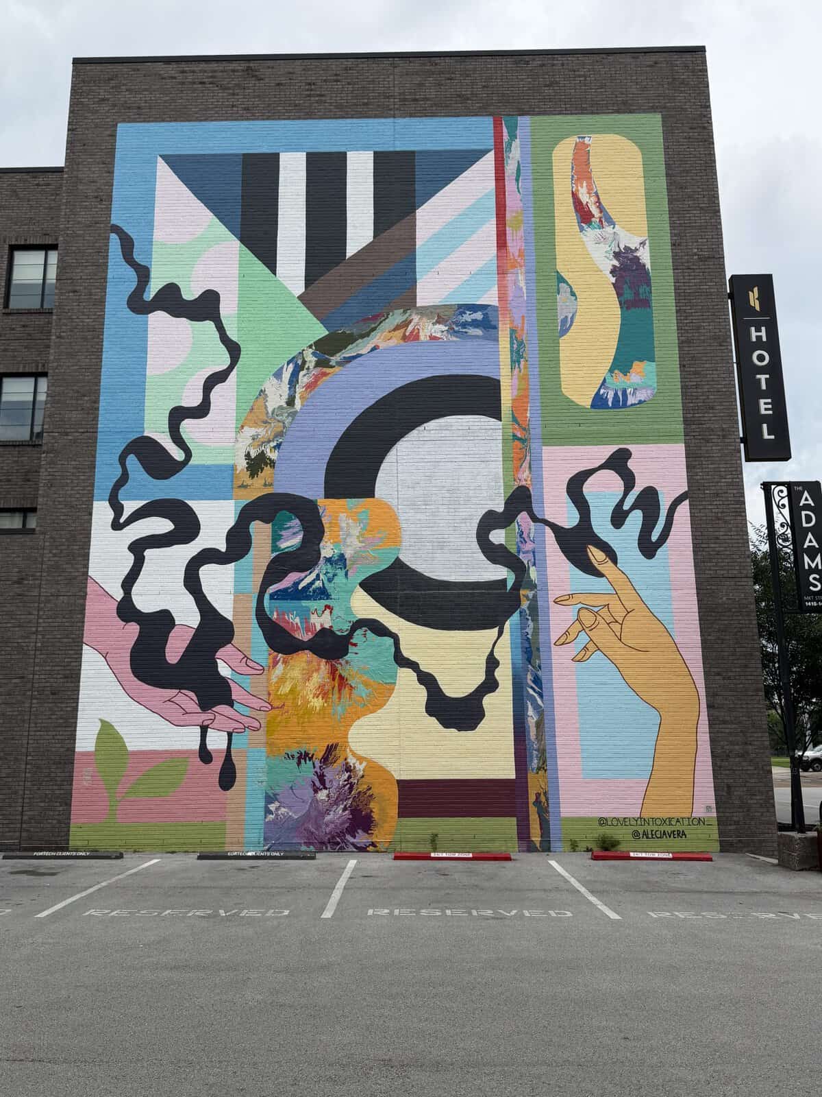 Large mural on brick wall featuring bright abstract shapes and a drawn hand reaching up, with a black squiggly line and colorful blocks around it.