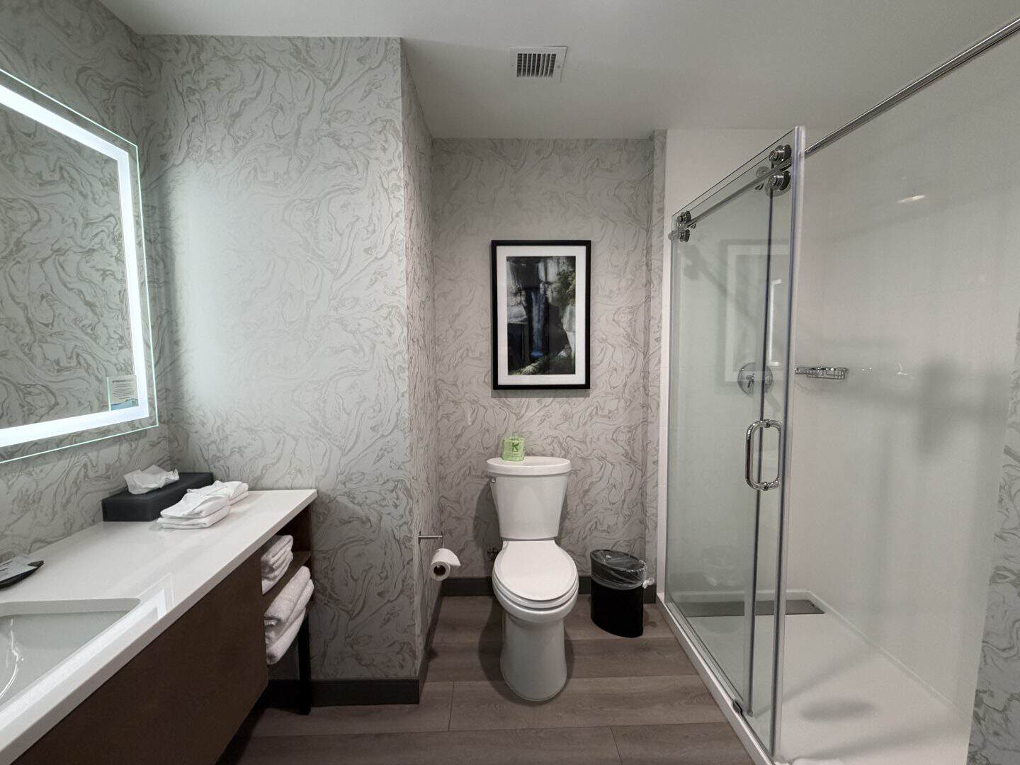 la_quinta_chattanooga_downtown_bathroom - Adventures with Carli Modern bathroom with marble-patterned walls, backlit mirror, white vanity, towels, toilet, framed art, and a glass shower enclosure.