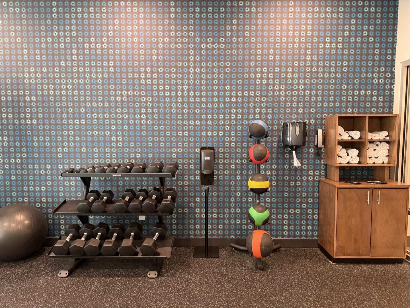 la_quinta_chattanooga_downtown_gym_weight_rack - Adventures with Carli La Quinta Chattanooga Downtown Fitness center with a blue patterned wall, dumbbell rack, stacked kettlebells, medicine balls on stands, and a wooden towel shelf nearby.