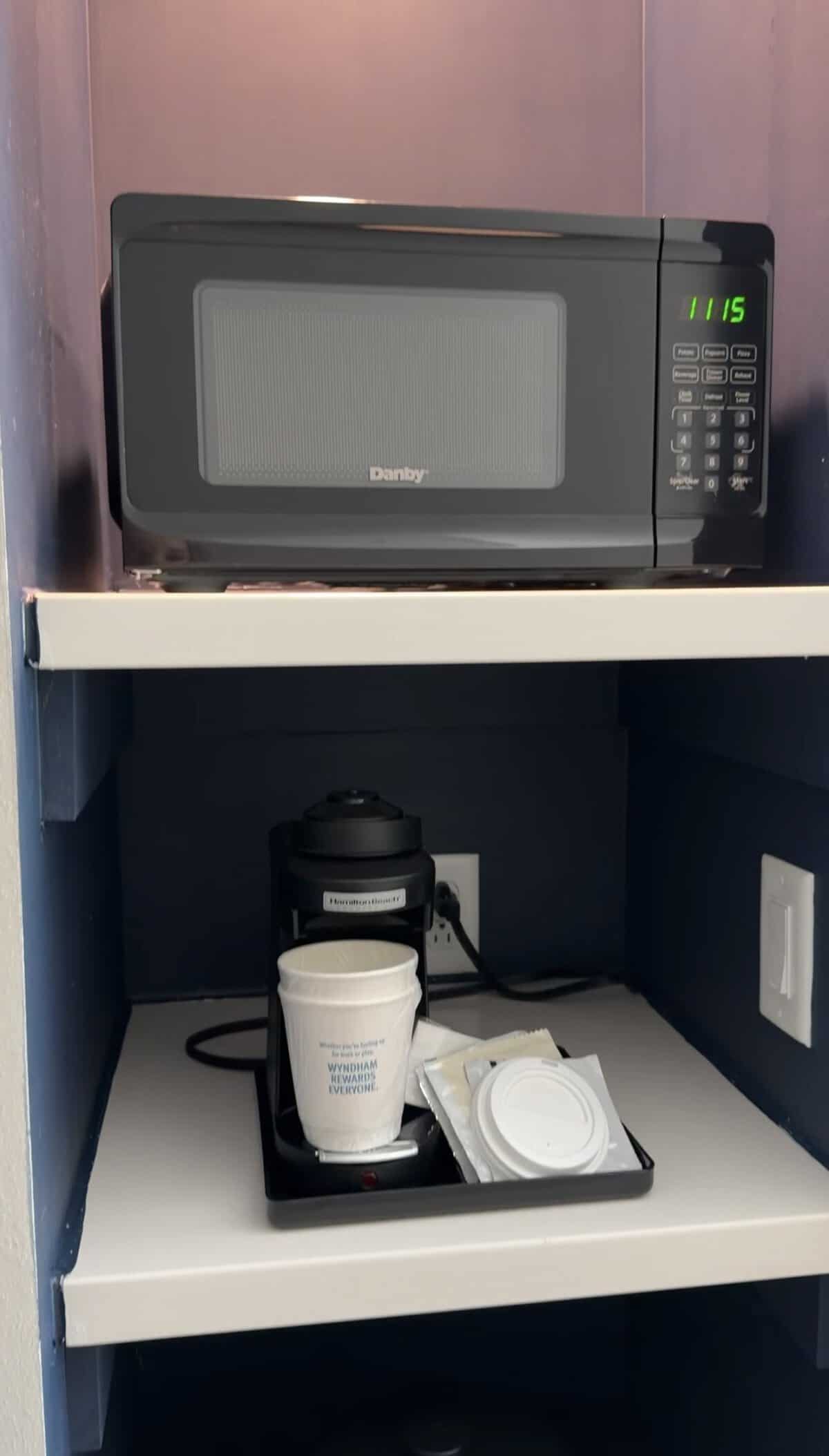 Screenshot - Adventures with Carli Microwave on a shelf above a compact coffee station with a coffee maker, cups, and napkins on the counter.