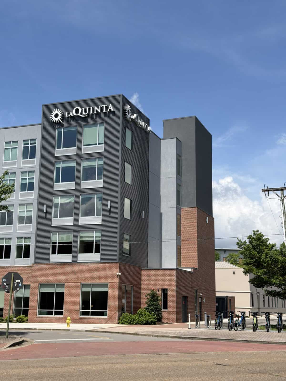 la_quinta_chattanooga_downtown_south - Adventures with Carli La Quinta Chattanooga Downtown hotel building with grey metal siding and brick base, bike rack and storefront along a red brick street beneath a blue sky of clouds-free day.
