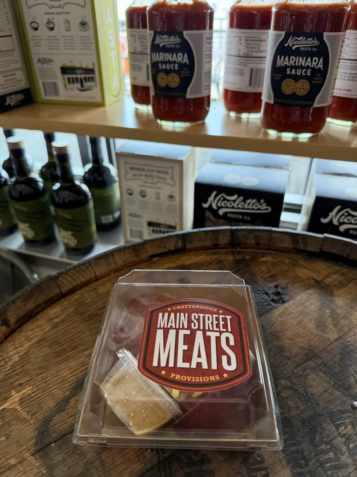 Plastic package labeled Main Street Meats Provisions sits on a wooden barrel table, with shelves of marinara sauce and pasta boxes in the background.