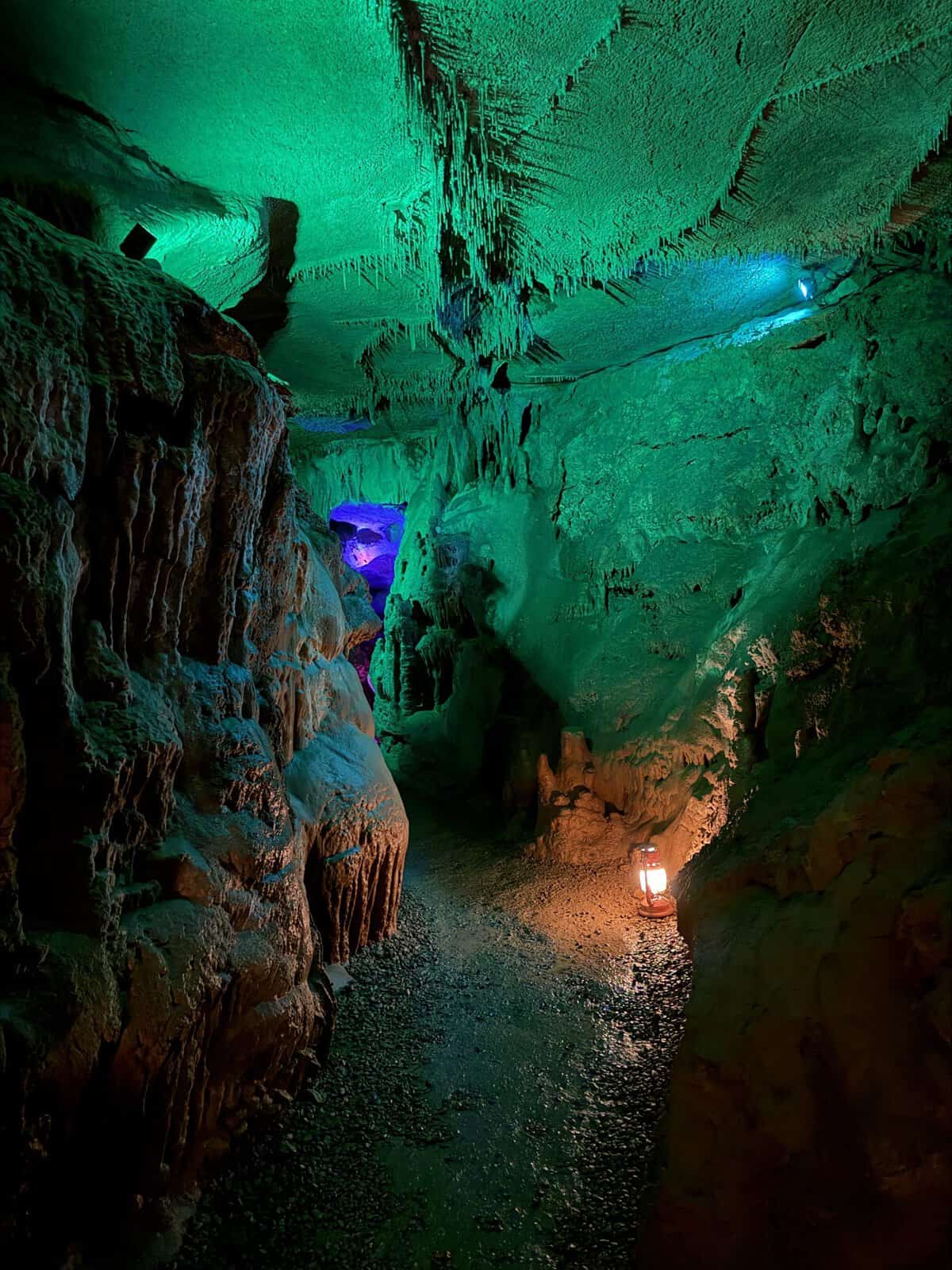 Cave interior with hanging stalactites, illuminated in green and blue, and a lantern on the rocky path.