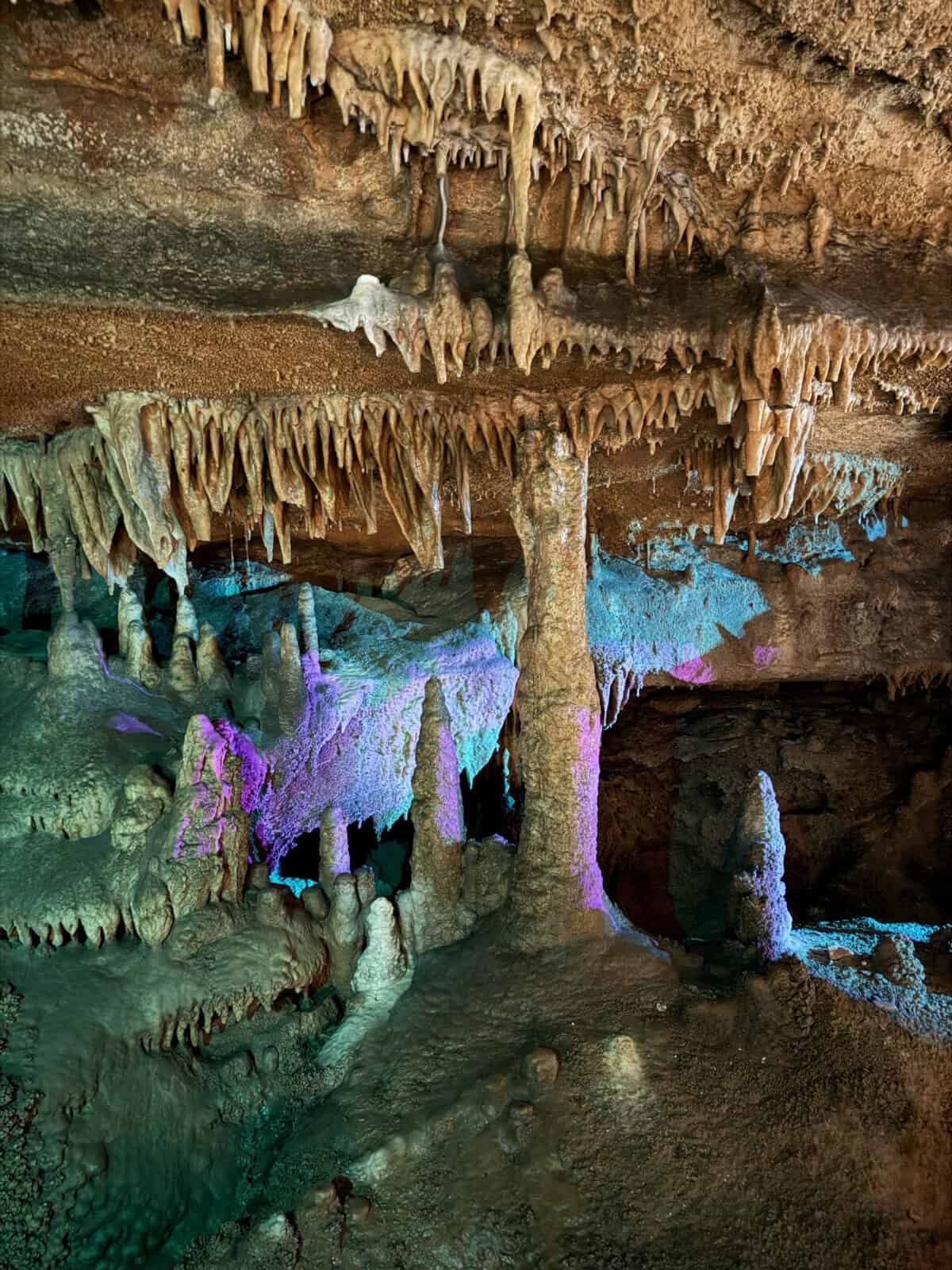Inside a dim limestone cave with numerous stalactites hanging from the ceiling and stalagmites forming pillars, lit by multicolored artificial lights.