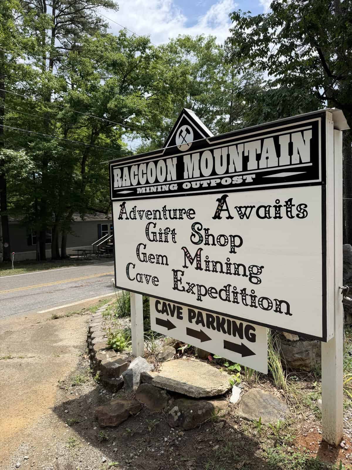 Sign for Raccoon Mountain Mining Outpost with listings: Adventure Awaits Gift Shop, Gem Mining, Cave Exploration, and parking arrows nearby.