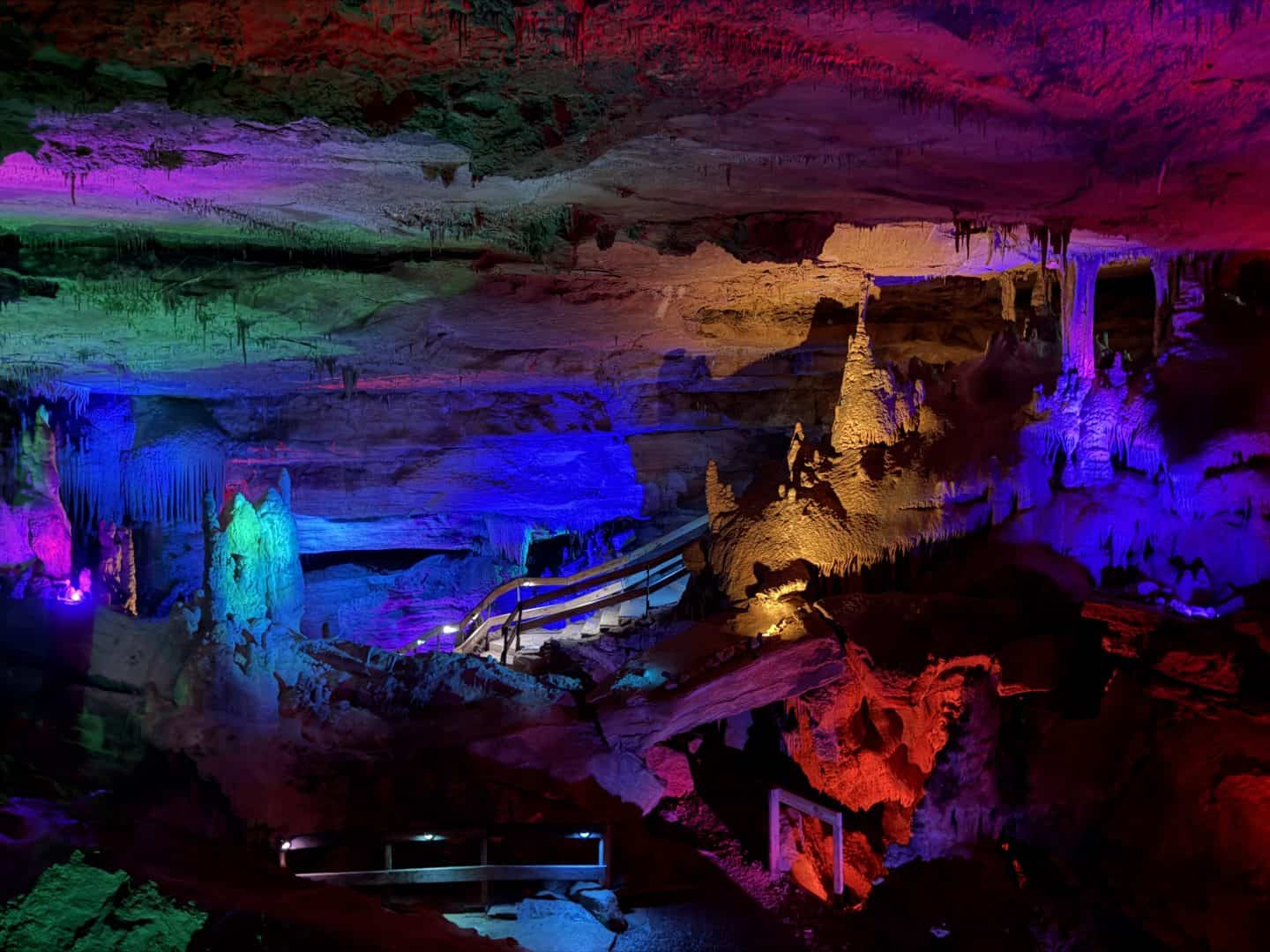 Underground cavern with stalactites illuminated by multicolored lights and a wooden railing path.