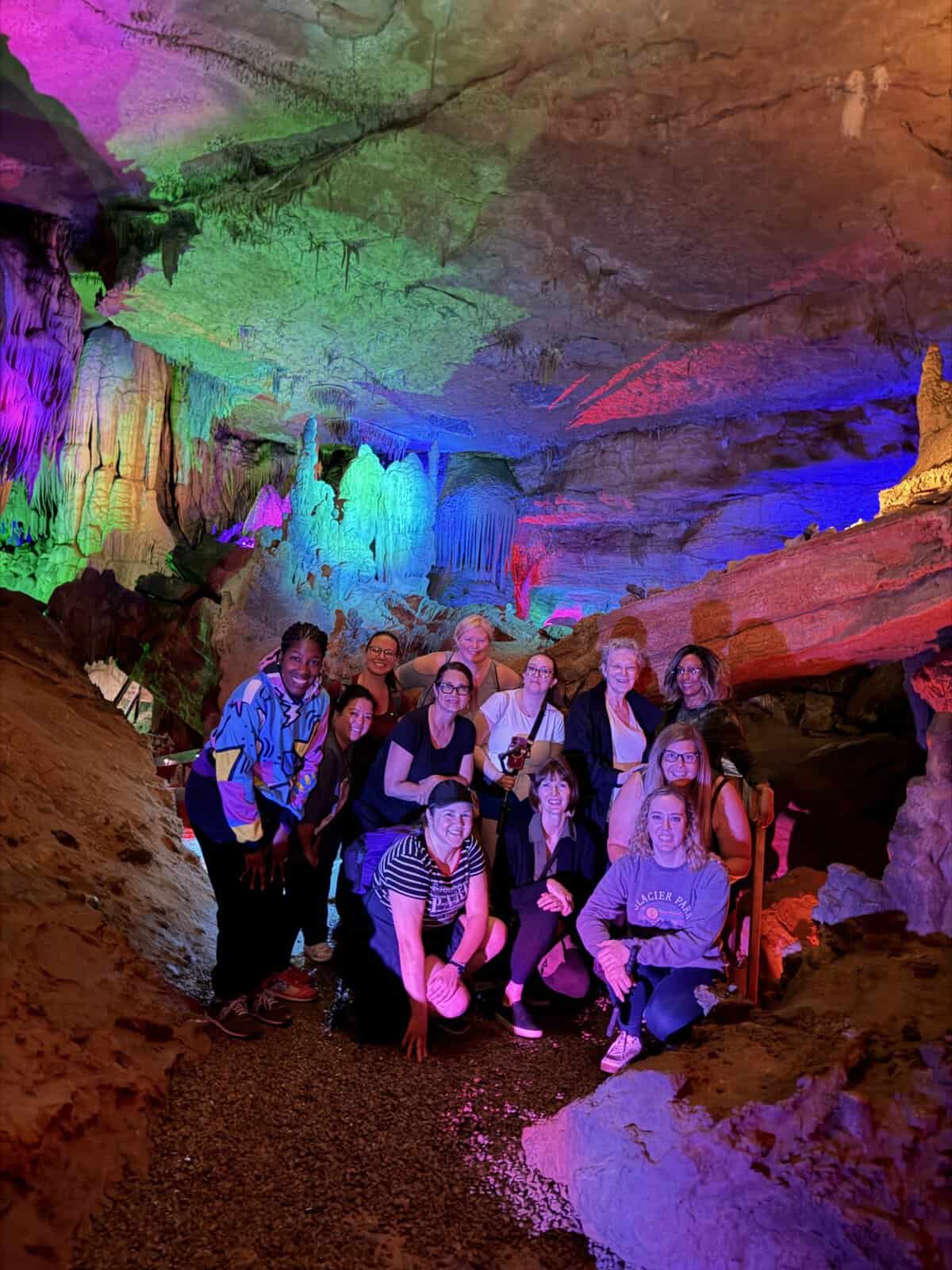 Group of women on Raccoon Mountain Caverns tours posing inside a colorful illuminated cave with stalagmites and rock formations behind them.