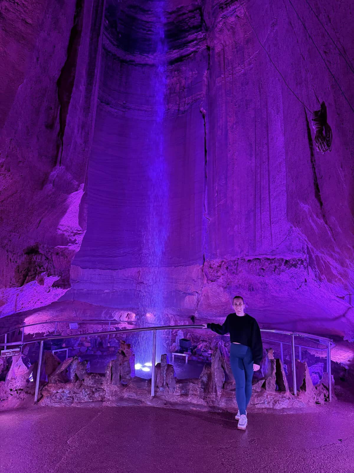 Carli standing by a metal railing inside a purple-lit cave, with waterfall in background