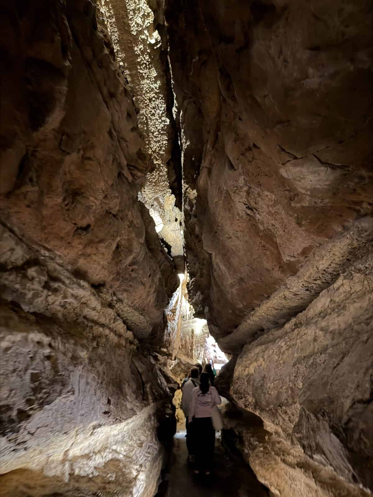People walking through a narrow cave passage with rough brown rock walls and light at the far end.