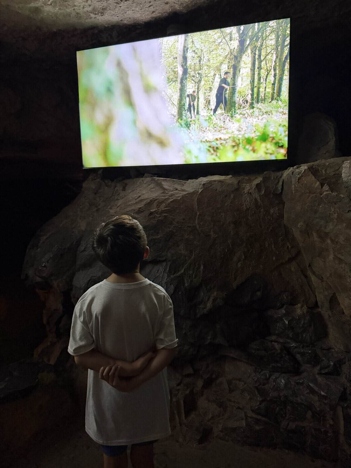 Boy in a white shirt stands in a dim cave, watching a large screen showing hikers in a sunlit forest.