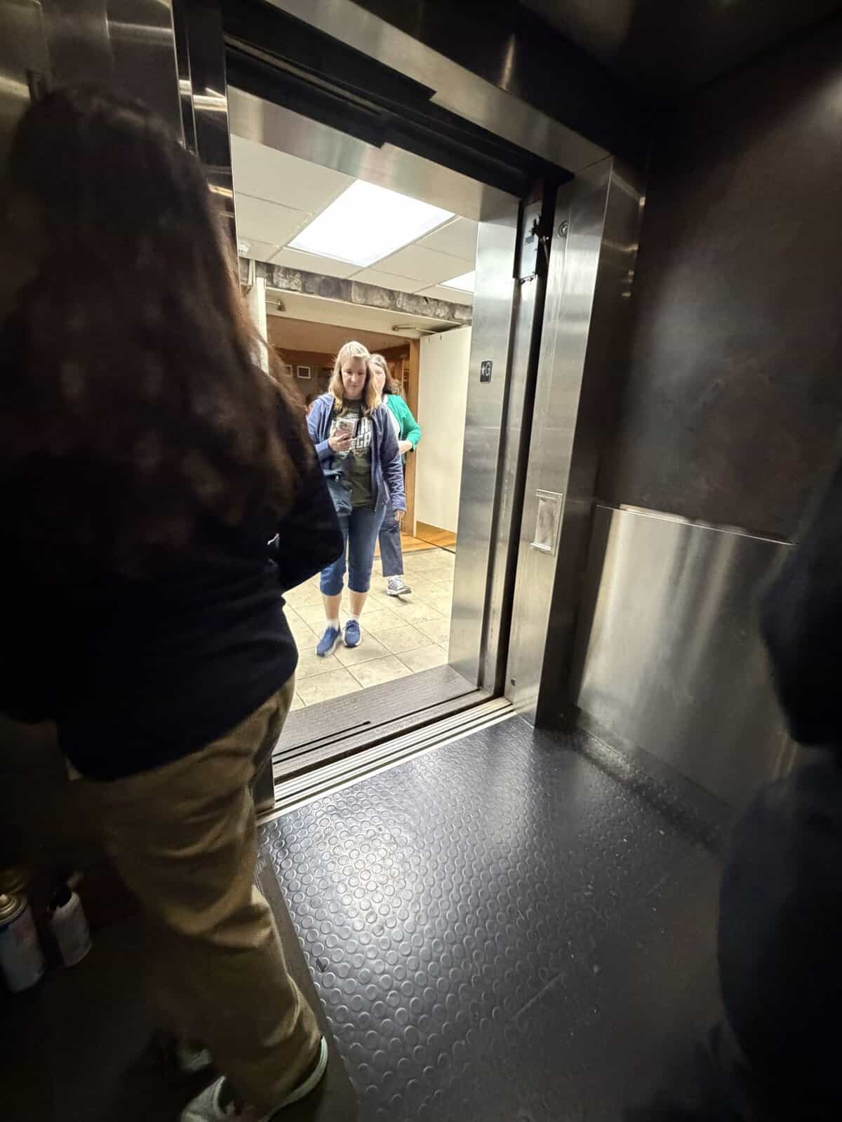 View from inside an open elevator as two people stand in the hallway outside; a young woman in a blue jacket looks at her phone while another person stands behind her.