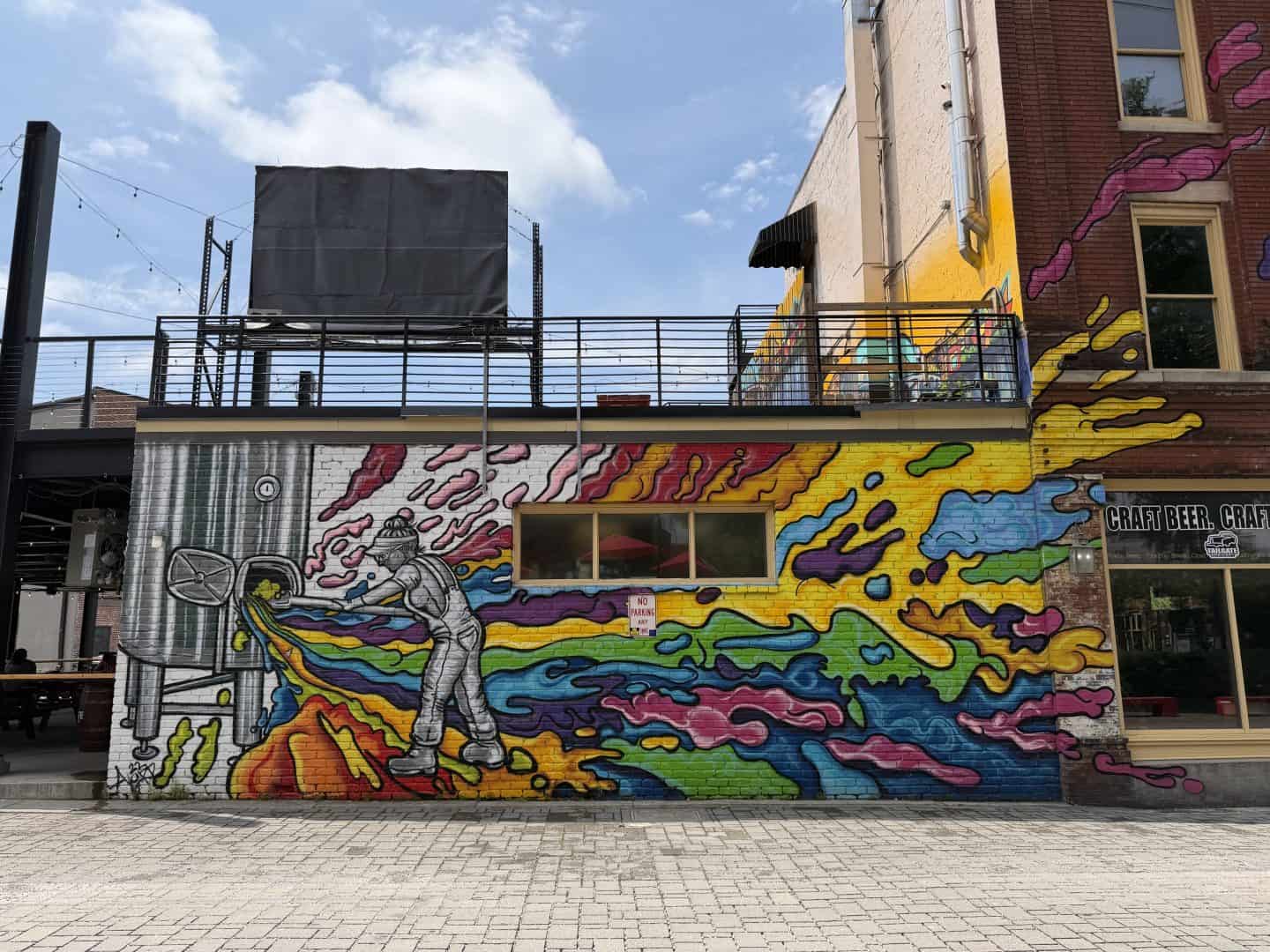Vibrant street mural on a brick wall: a person in overalls sprays paint, creating rainbow-colored, flowing shapes across the wall beneath a railing and rooftop area.