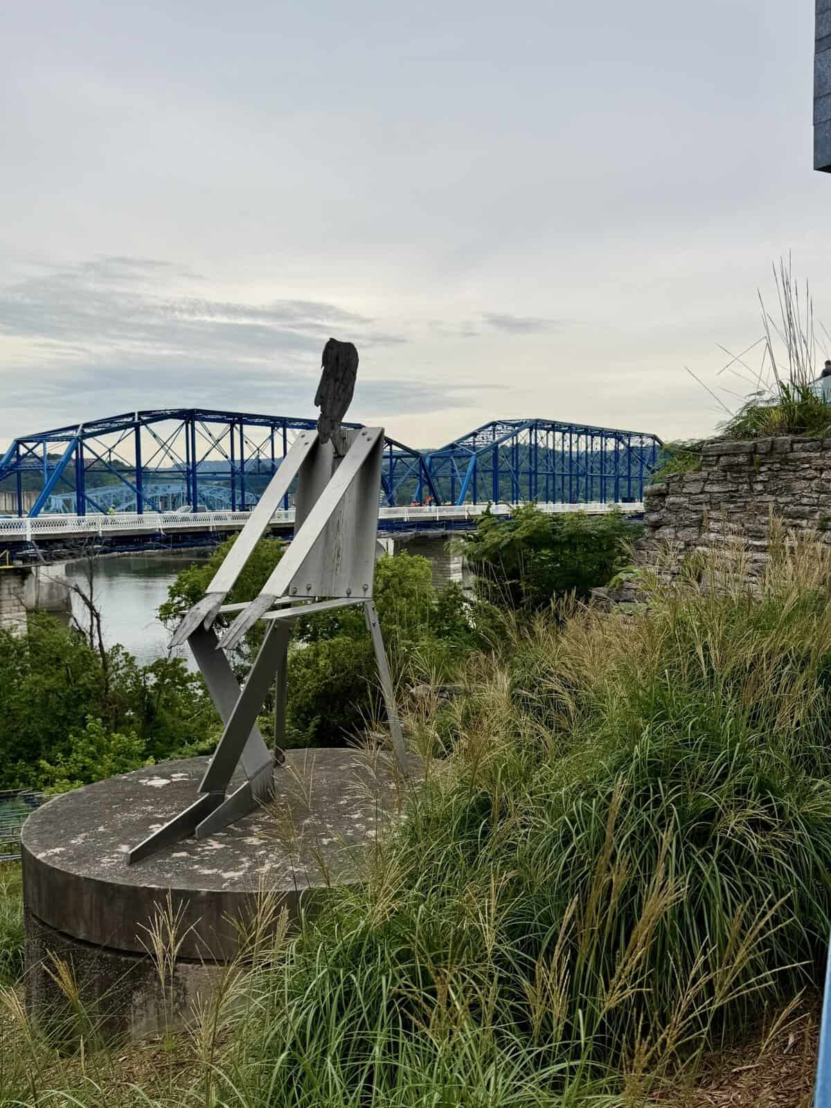 Modern metal sculpture on a circular concrete base with a blue truss bridge across a river in the distance, grassy foreground.