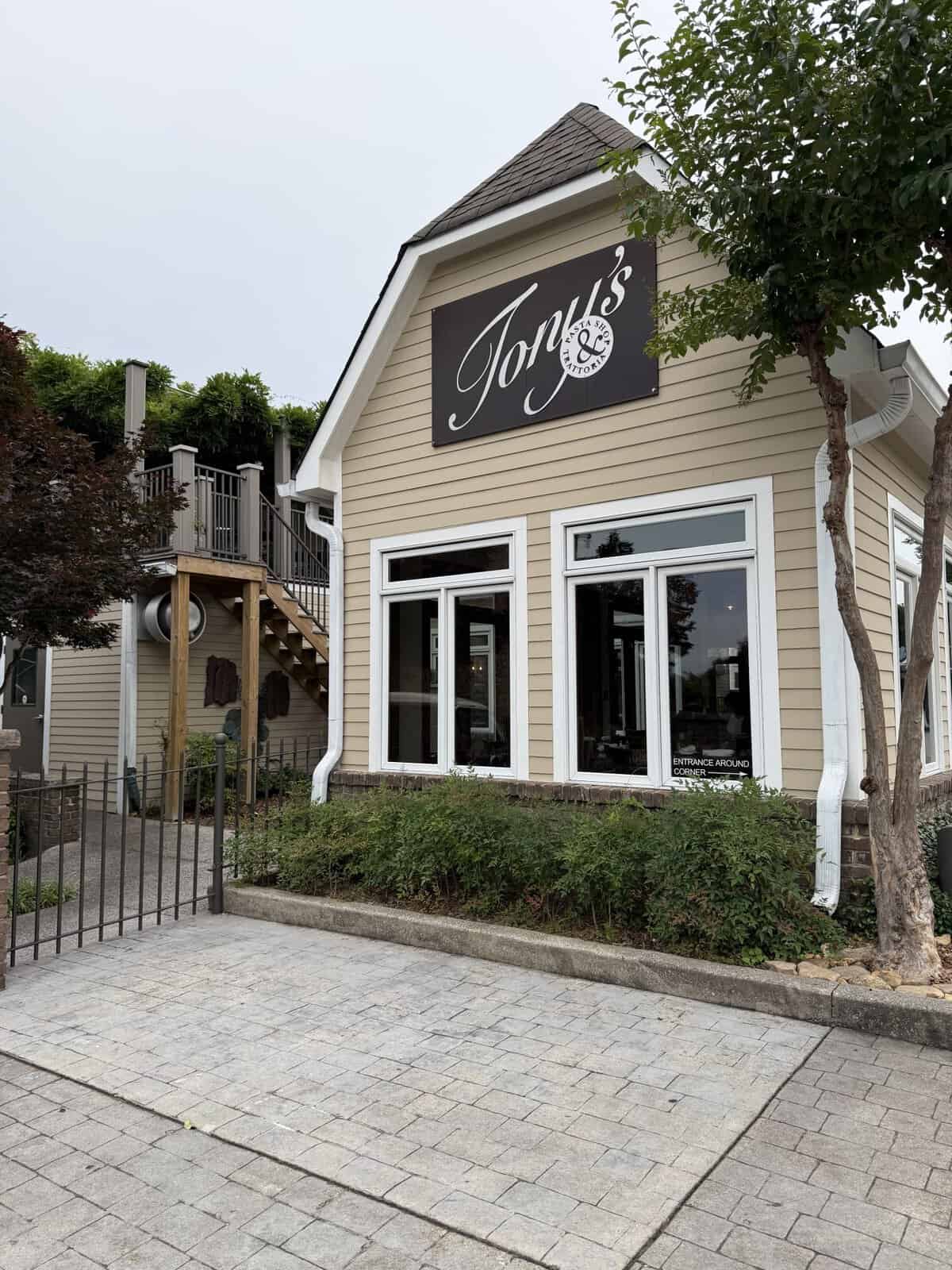 Restaurant with a large black sign reading'Tony's' mounted on beige siding, white-framed windows, and a small tree nearby.