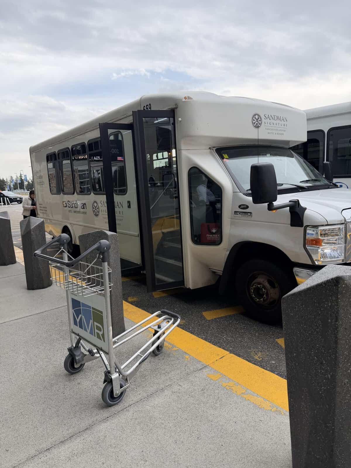 White Sandman Signature shuttle bus with its front door open, parked at a curbside in a parking lot; a luggage cart is in the foreground.