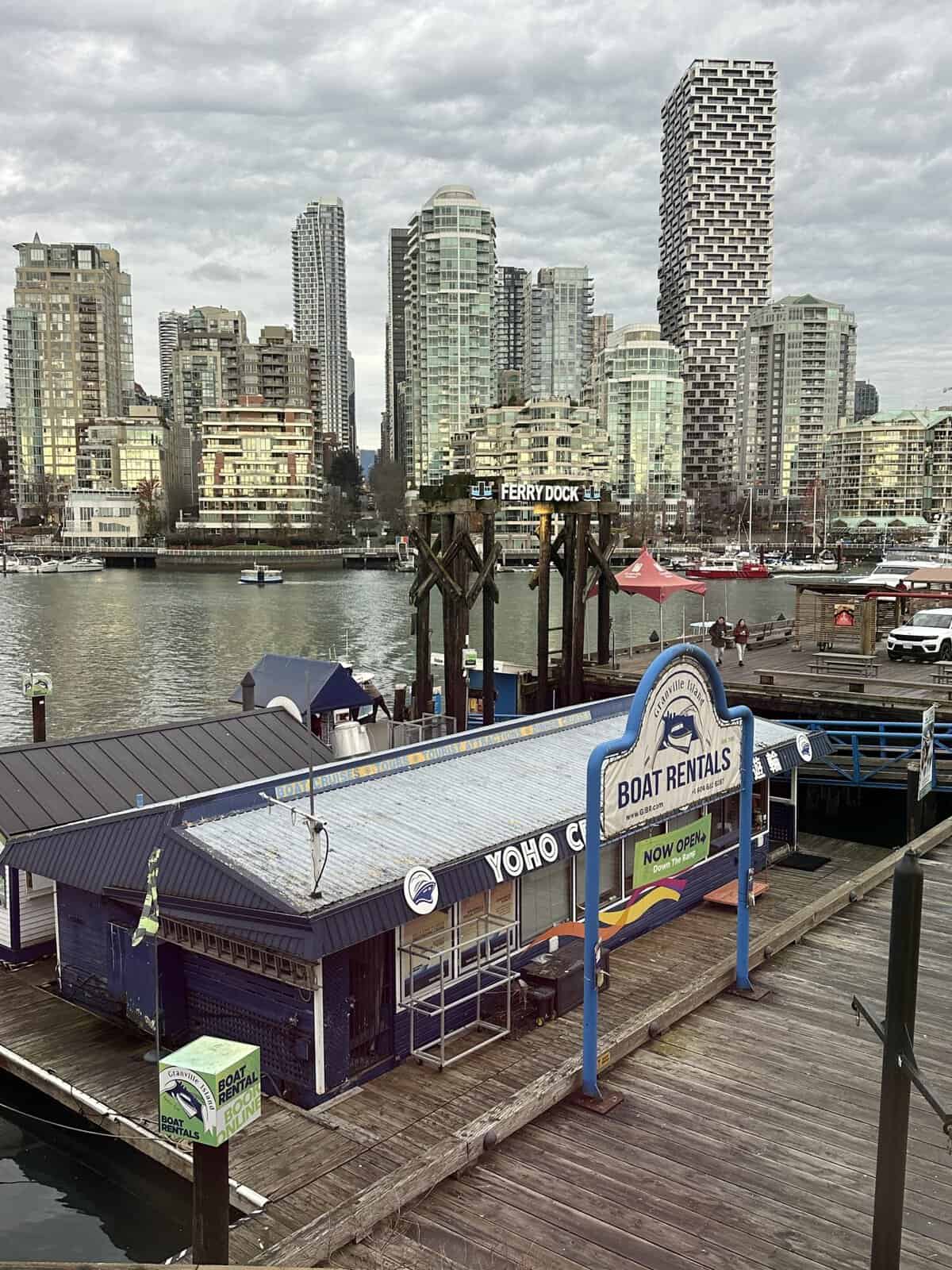 Wooden dockside boat rental shop with blue facade and signs reading'Boat Rentals' and 'Now Open', with a city marina and high-rise buildings in the background.