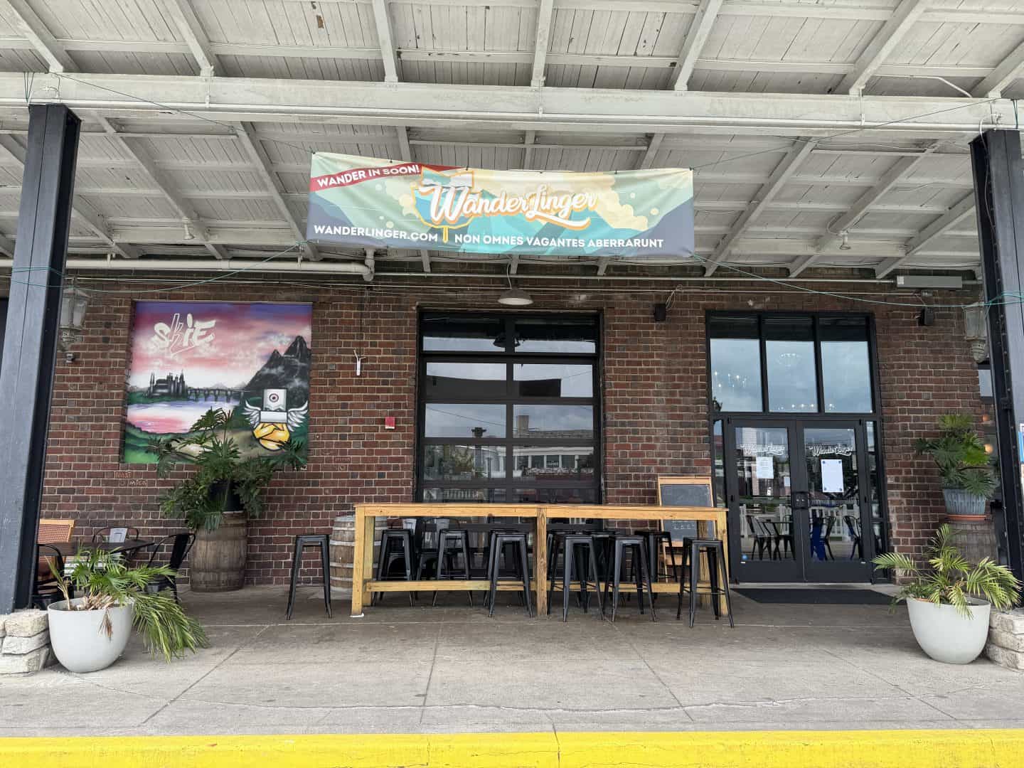 exterior of wanderlinger brewery.  brick wall with black trim and large glass windows and doors.  banner hanging from ceiling.  tall wood table in center with black backless stools.
