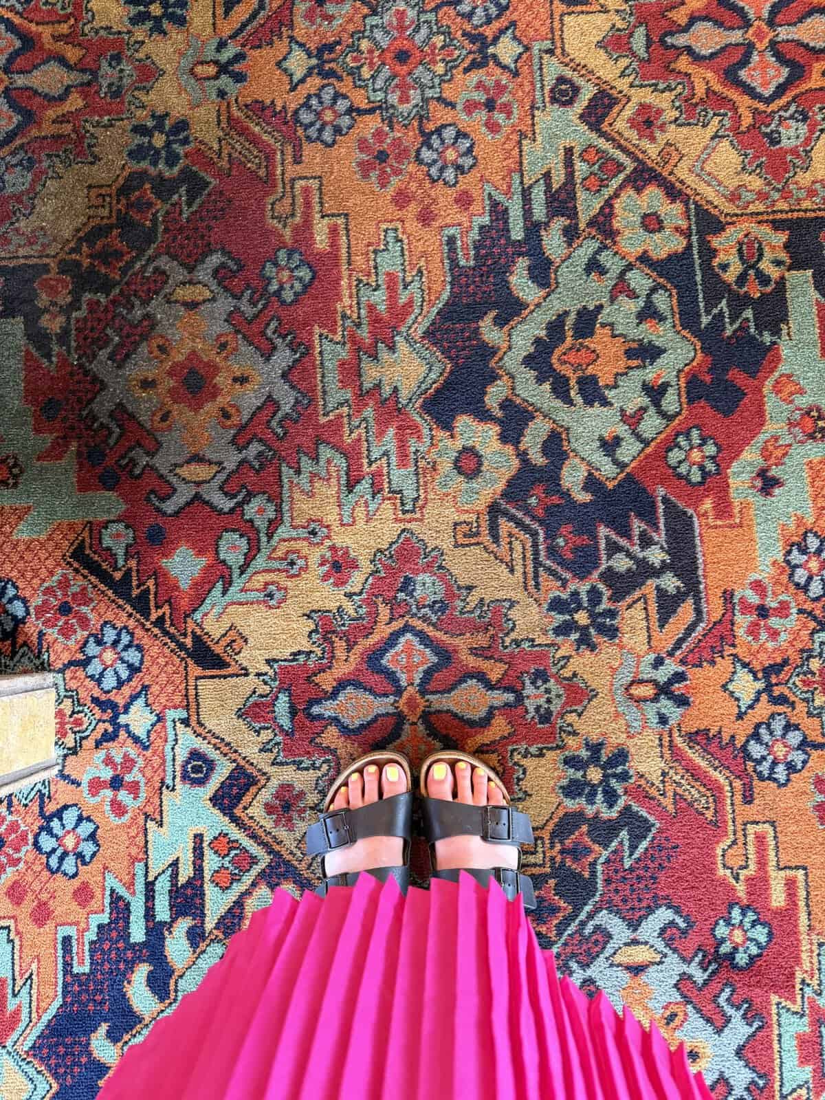 Feet in black sandals on a colorful patterned rug, with a bright pink pleated dress visible.