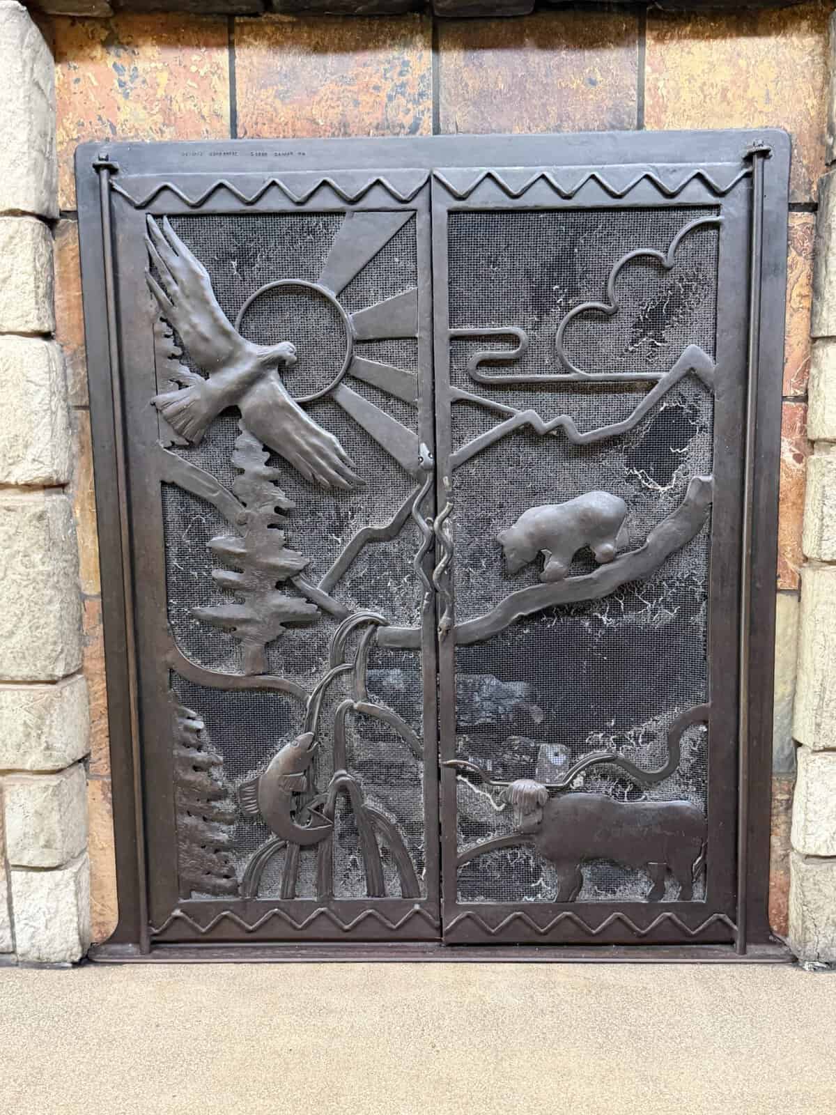 Decorative metal gate with nature relief: a flying bird, sun rays, mountains, pine tree, and a bear on a slope in two panels.