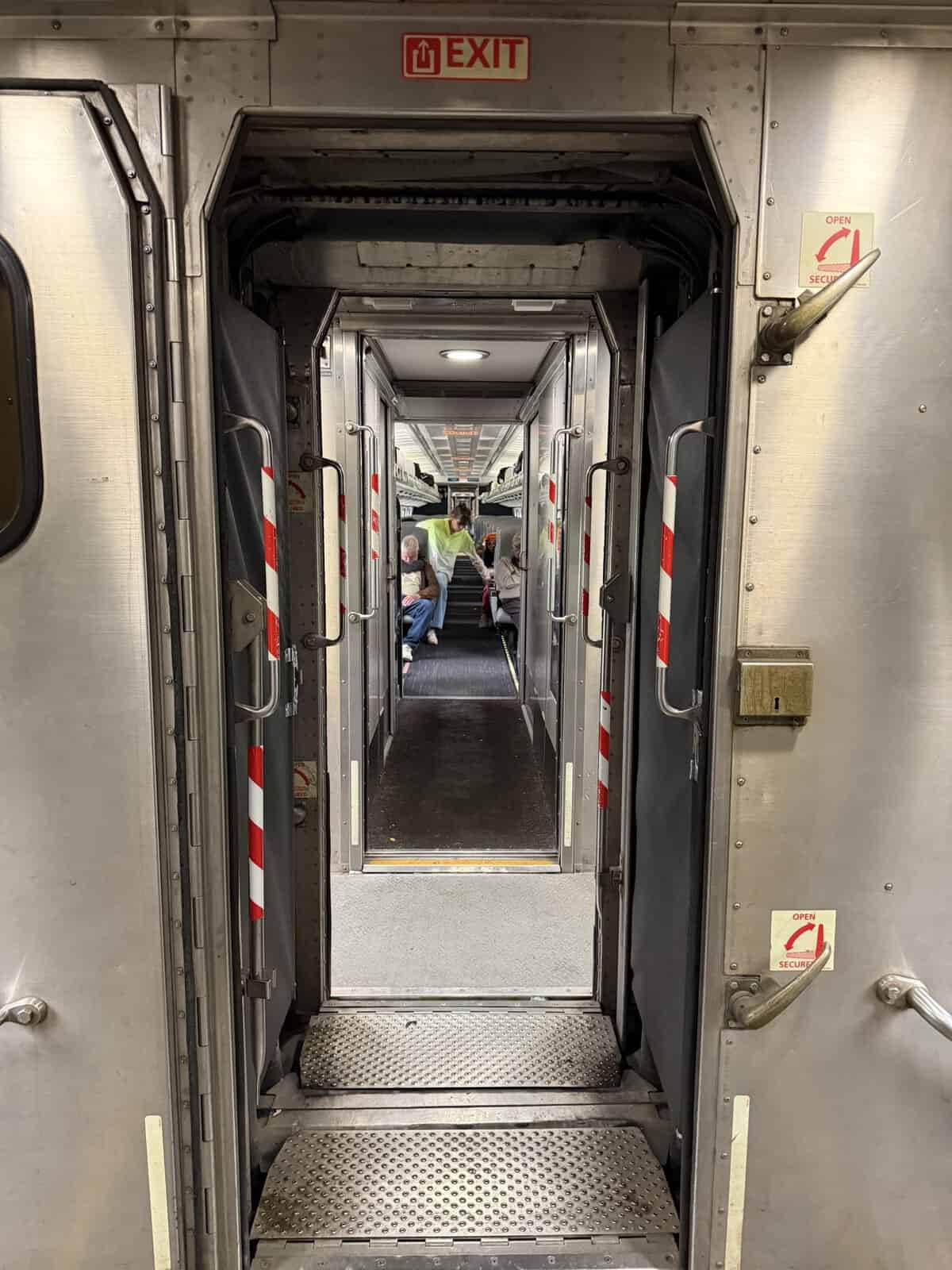 Industrial train doorway looking into a dimly lit commuter car, red-and-white striped barriers along the frame and passengers seated in the background