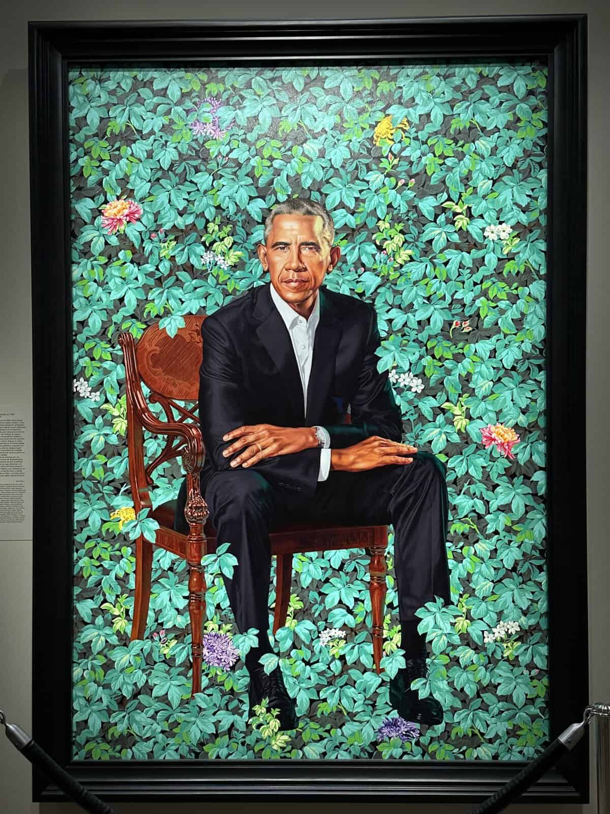 Portrait of Barack Obama seated in a black suit on a carved wooden chair, surrounded by dense teal ivy and colorful flowers.