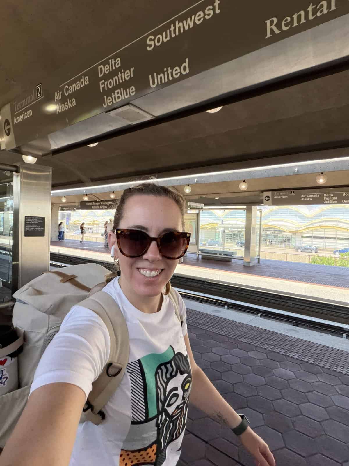 selfie of Carli on a train platform with overhead signs listing airline terminals and destinations.