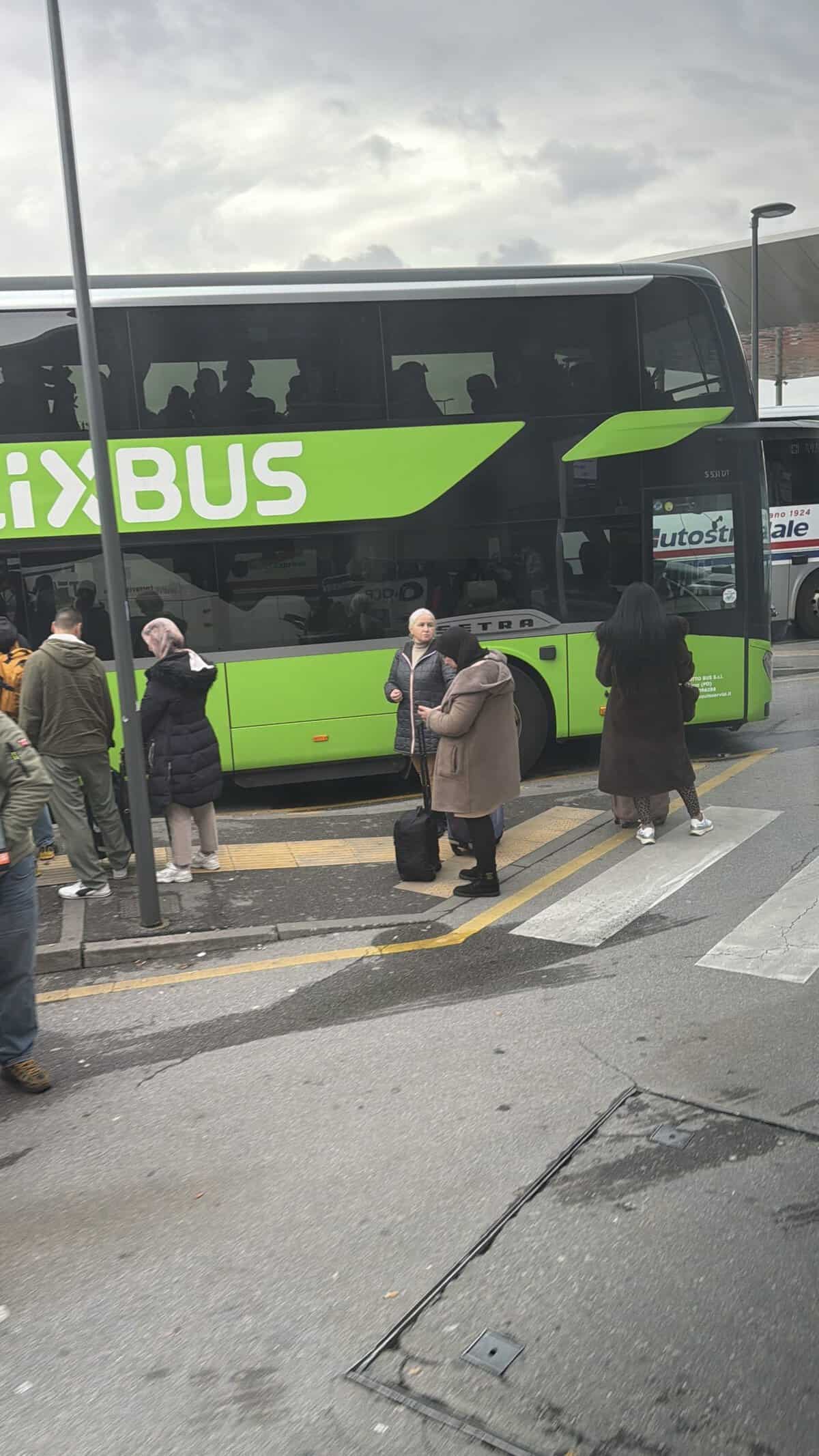 Bright green and black double-decker bus with passengers inside, stopped at a curb as people wait to board at a bus station.