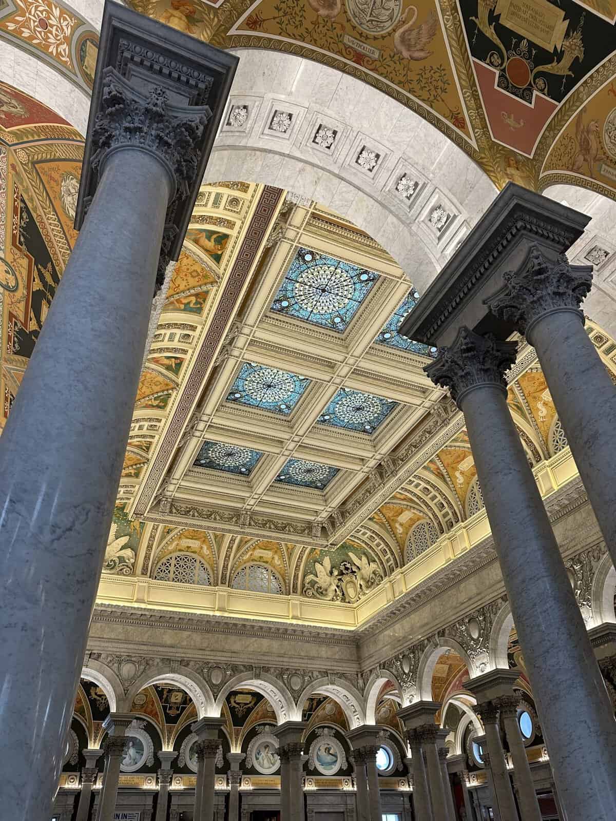 Interior of a grand historic atrium with tall marble columns supporting decorated arches and a colorful domed ceiling with mosaics and frescoes.