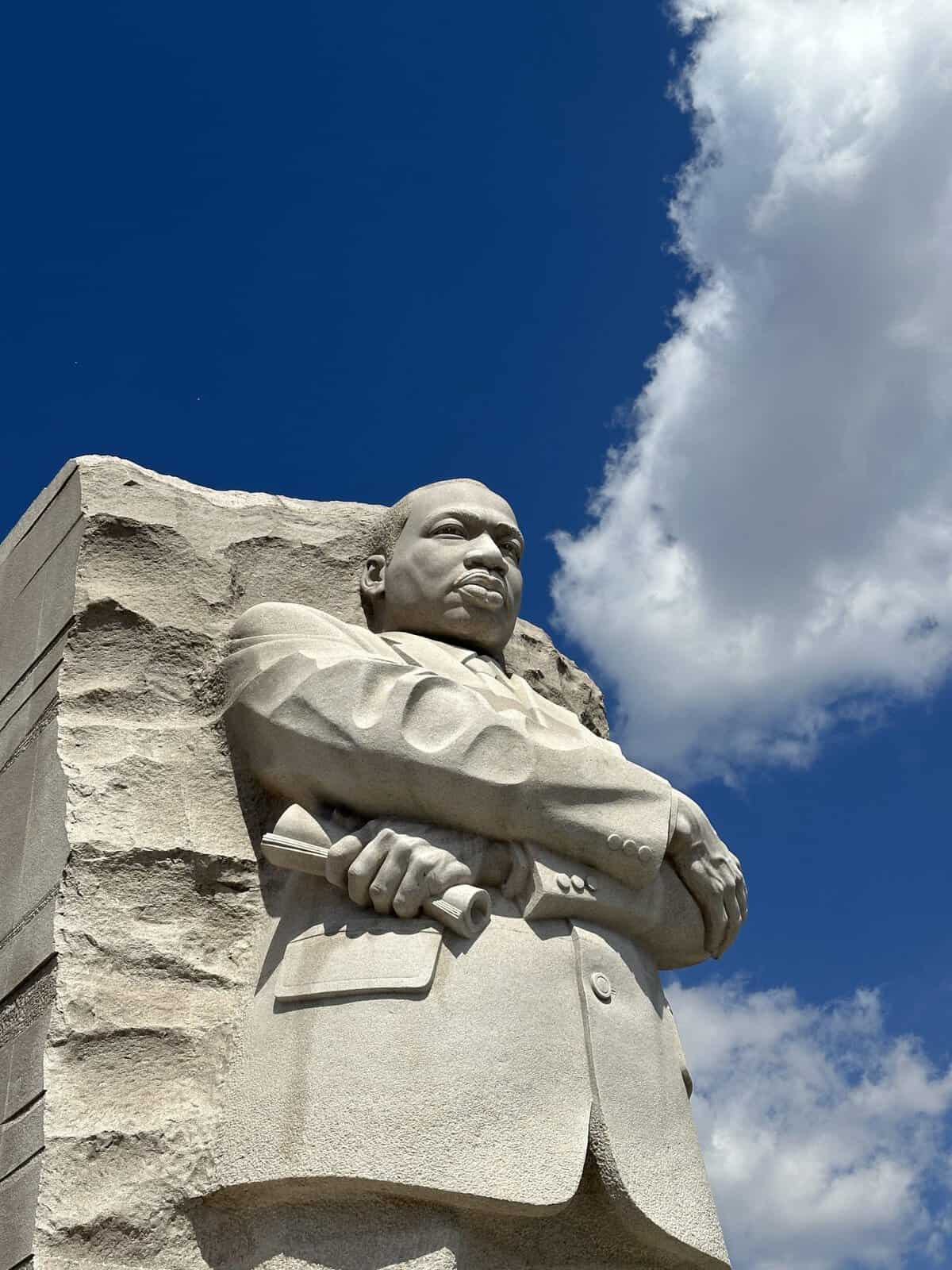 Stone sculpture of Martin Luther King JR gripping a rolled document, carved from granite with a blue sky and clouds behind him.