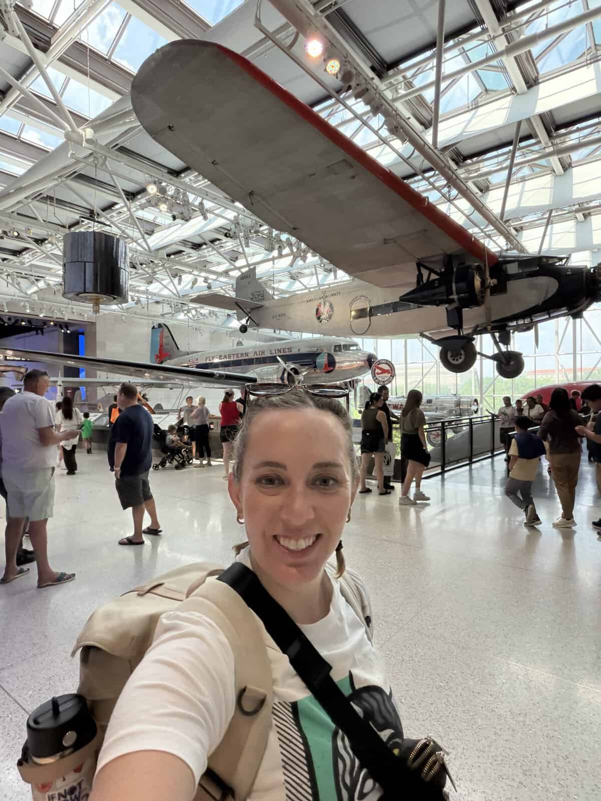Carli taking a selfie inside an aviation museum, with vintage aircraft suspended from the ceiling and visitors in the background.